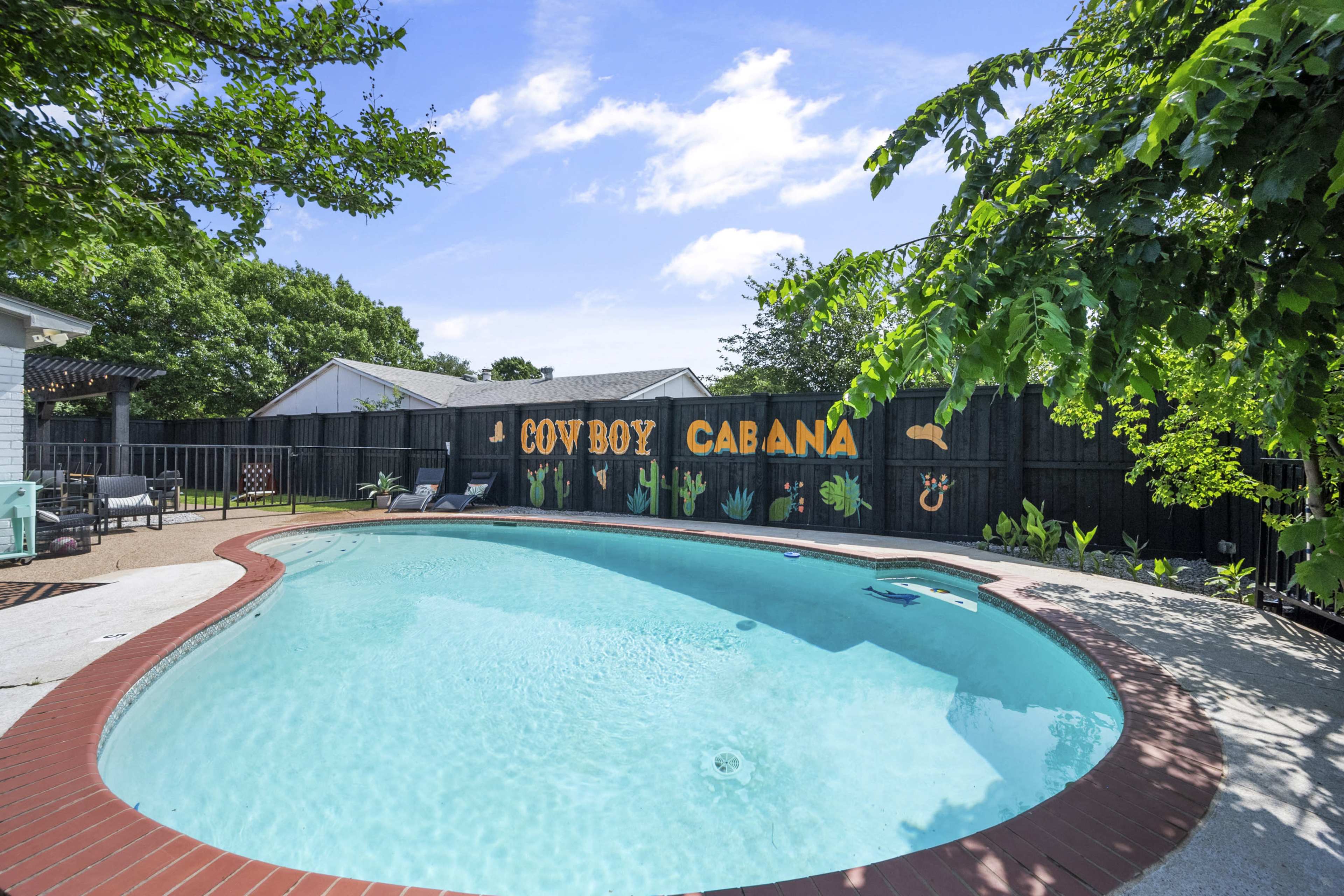 The image shows a swimming pool surrounded by greenery and decorated with a sign that reads "COWBOY CABANA" against a wooden fence.