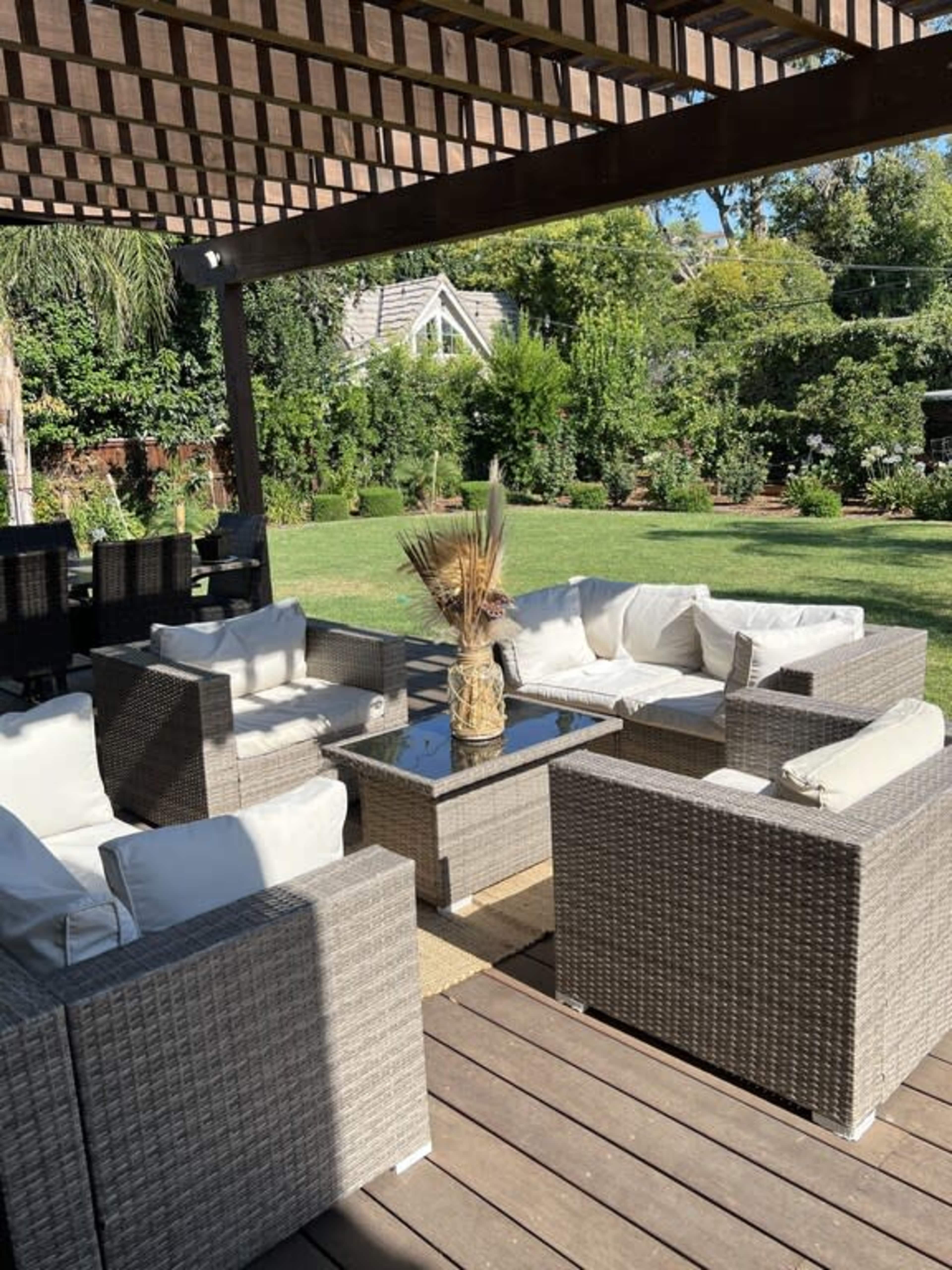 A patio seating area features wicker furniture with white cushions arranged around a glass-topped table, set beneath a wooden pergola in a landscaped yard.