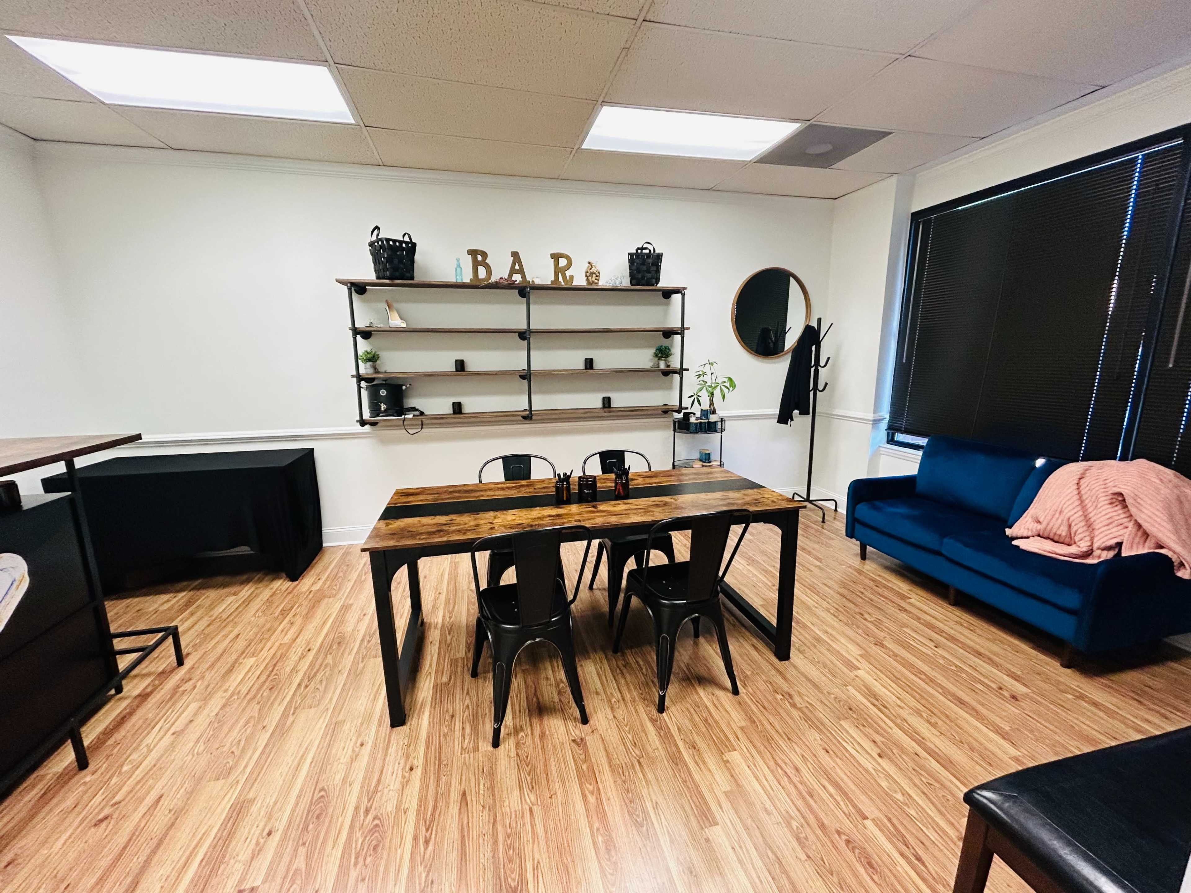 A modern office space featuring a wooden dining table surrounded by black chairs, a blue couch, and a shelving unit with decor items against a white wall.