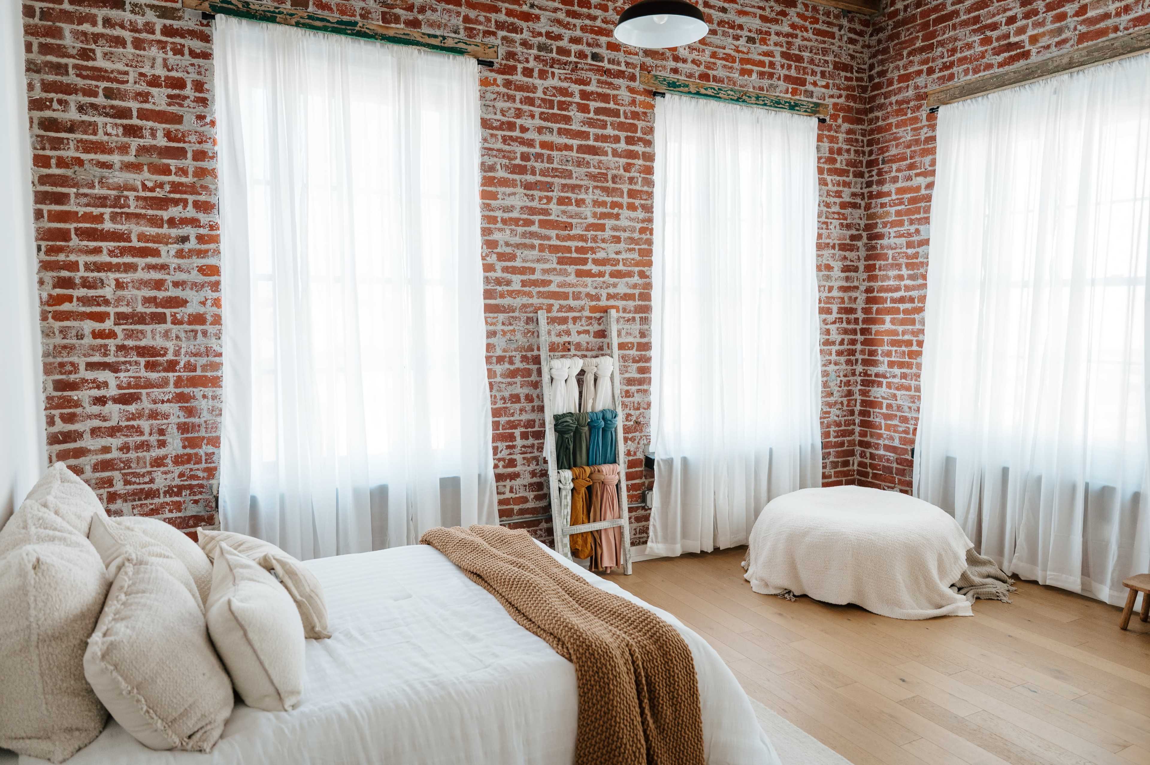 The image shows a bedroom with exposed brick walls, large windows draped in sheer white curtains, and a simple bed arrangement.