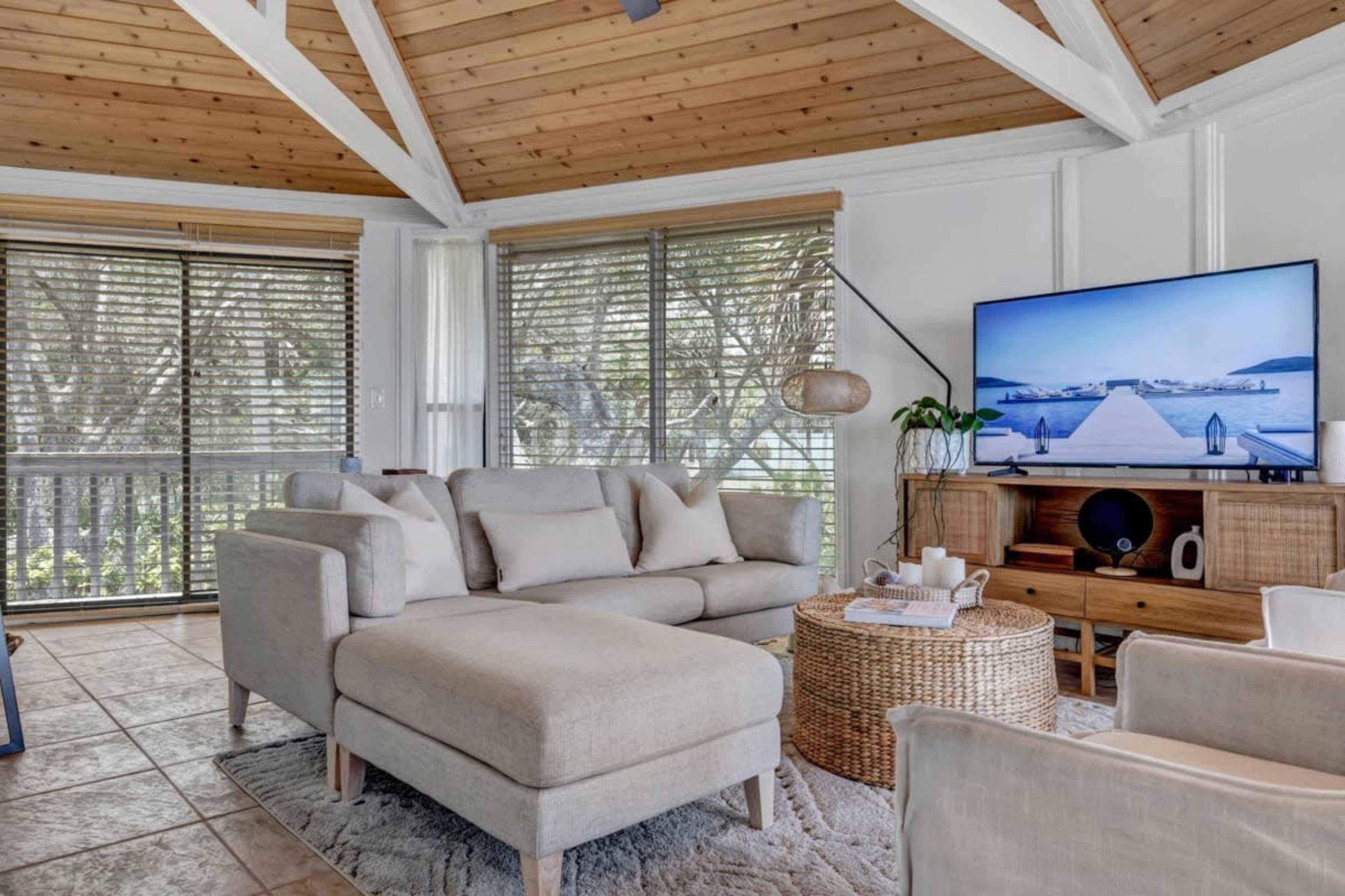 A spacious living room features a light-colored sectional sofa, a round wicker coffee table, and a large television mounted on a white wall, all set against wooden beams and large windows with blinds.