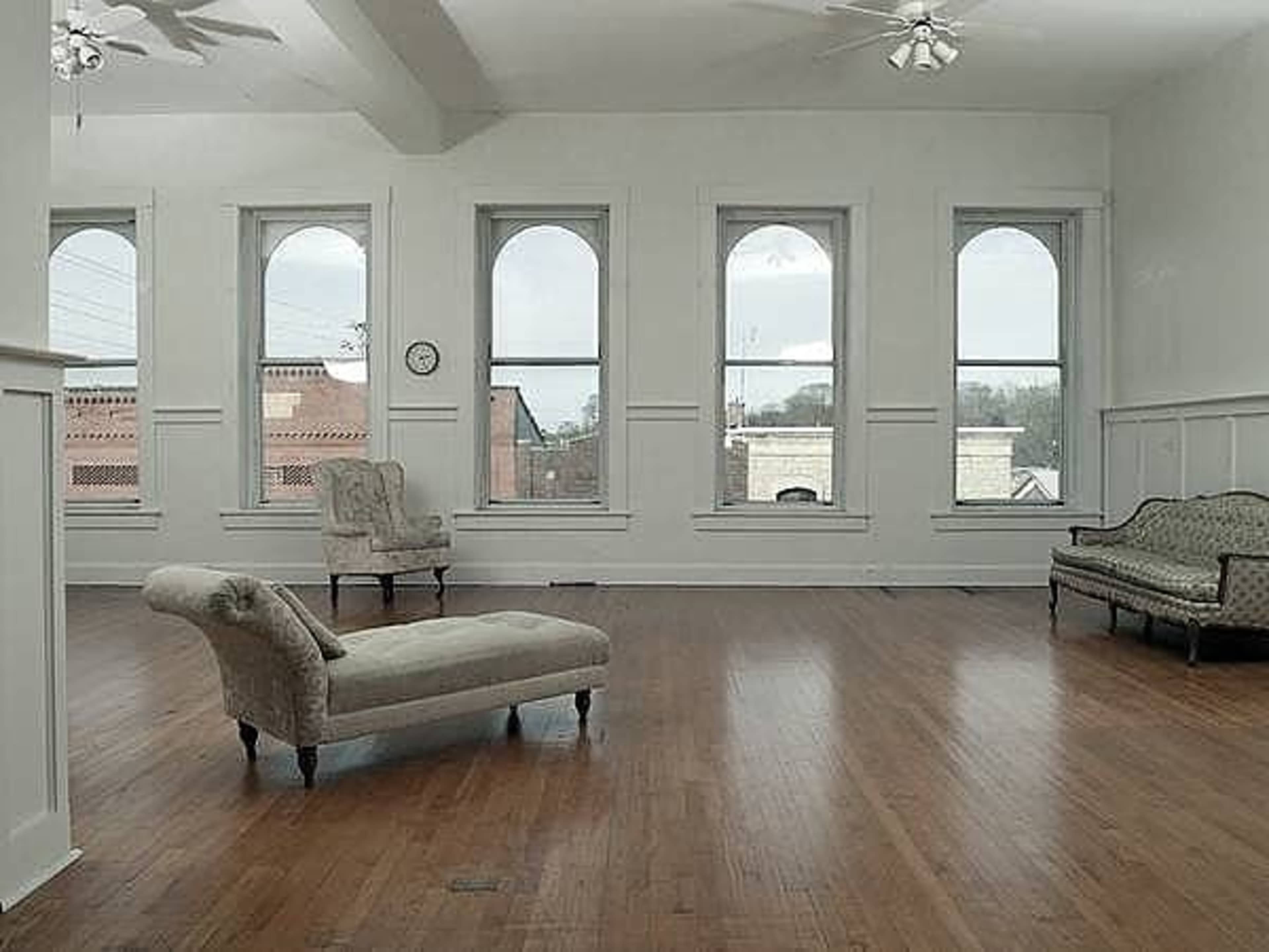 A spacious, light-filled room with large arched windows, hardwood flooring, and two upholstered chairs placed against the walls.