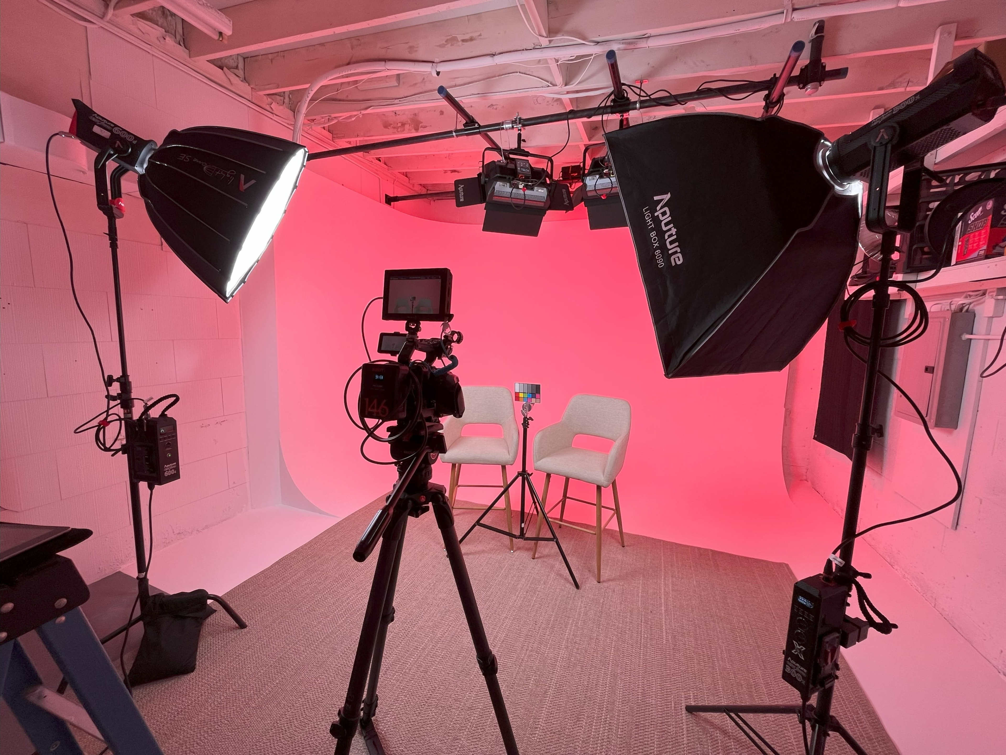 A photography setup featuring two empty chairs positioned in front of a pink backdrop, with studio lights and a camera on a tripod arranged around the scene.