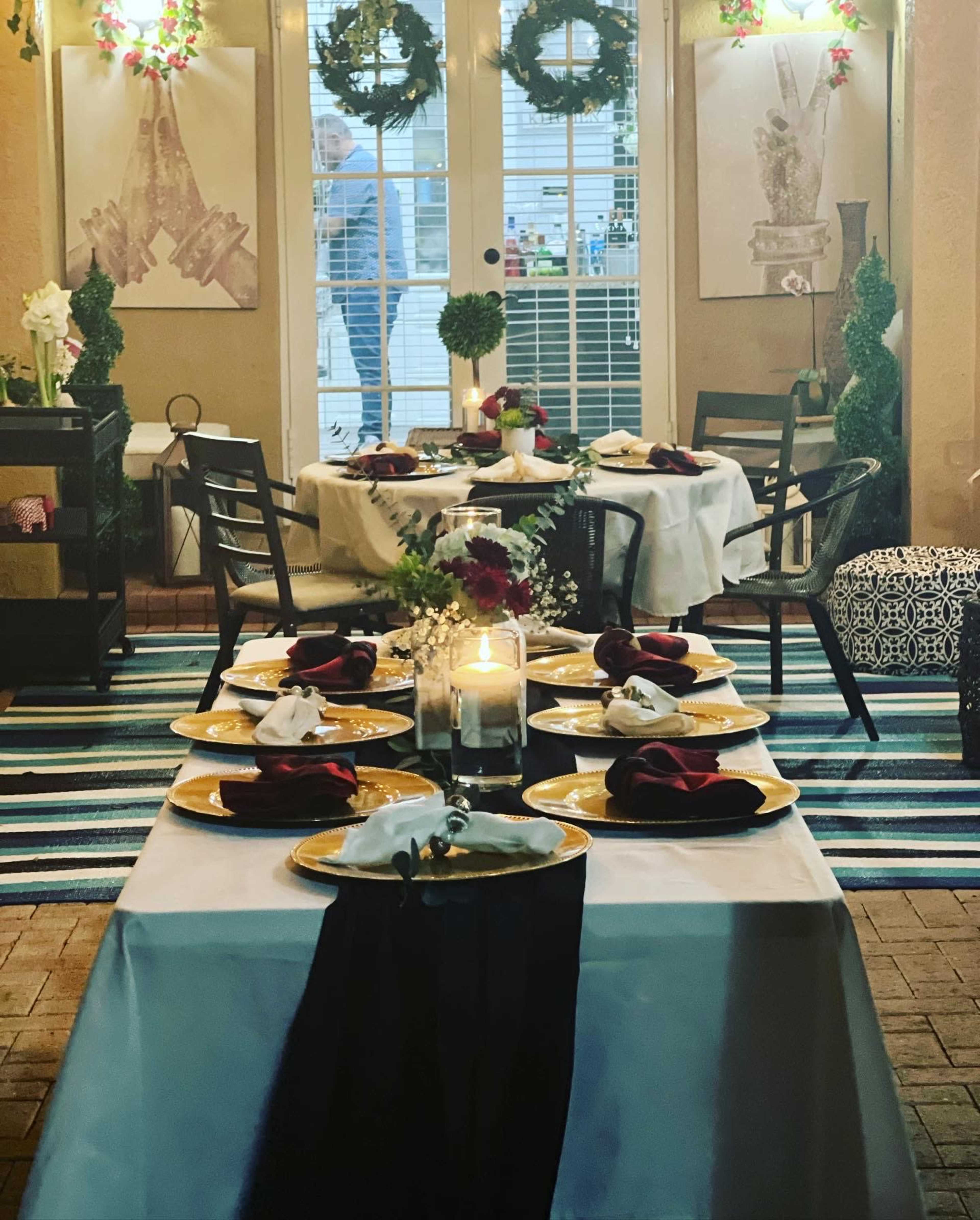 A dining setup featuring neatly arranged tables with white tablecloths, black runners, and gold dinnerware, surrounded by decorated walls and green wreaths.