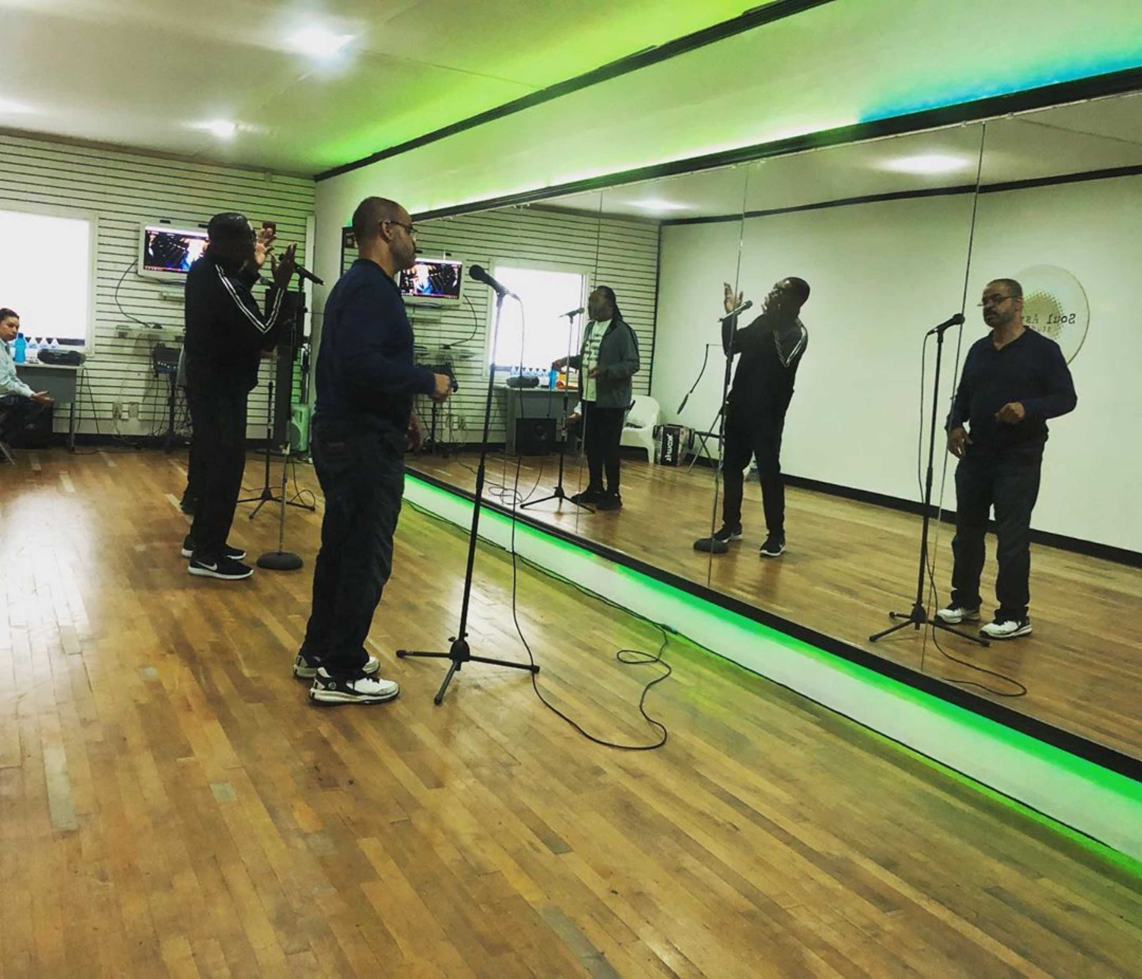 Two men are singing into microphones in a rehearsal studio with mirrors and green lighting.