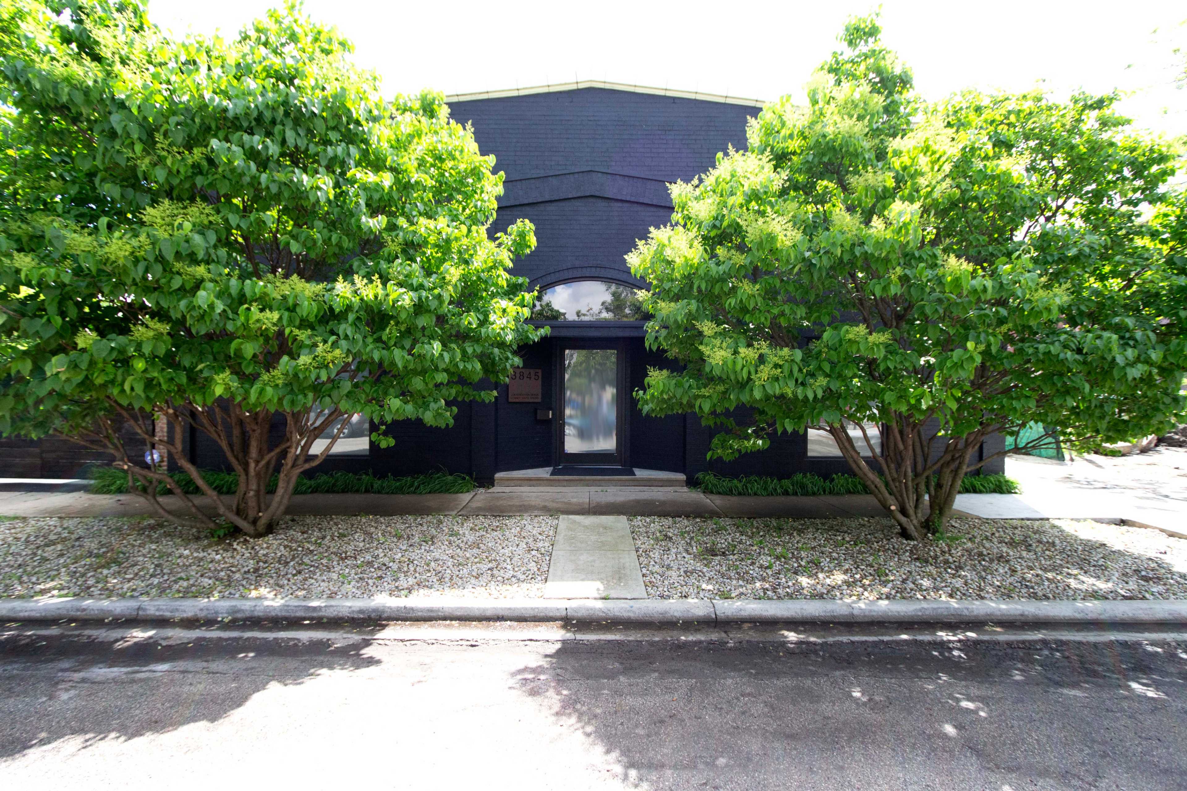 A modern black building framed by two green trees, with a clear pathway leading to the entrance.
