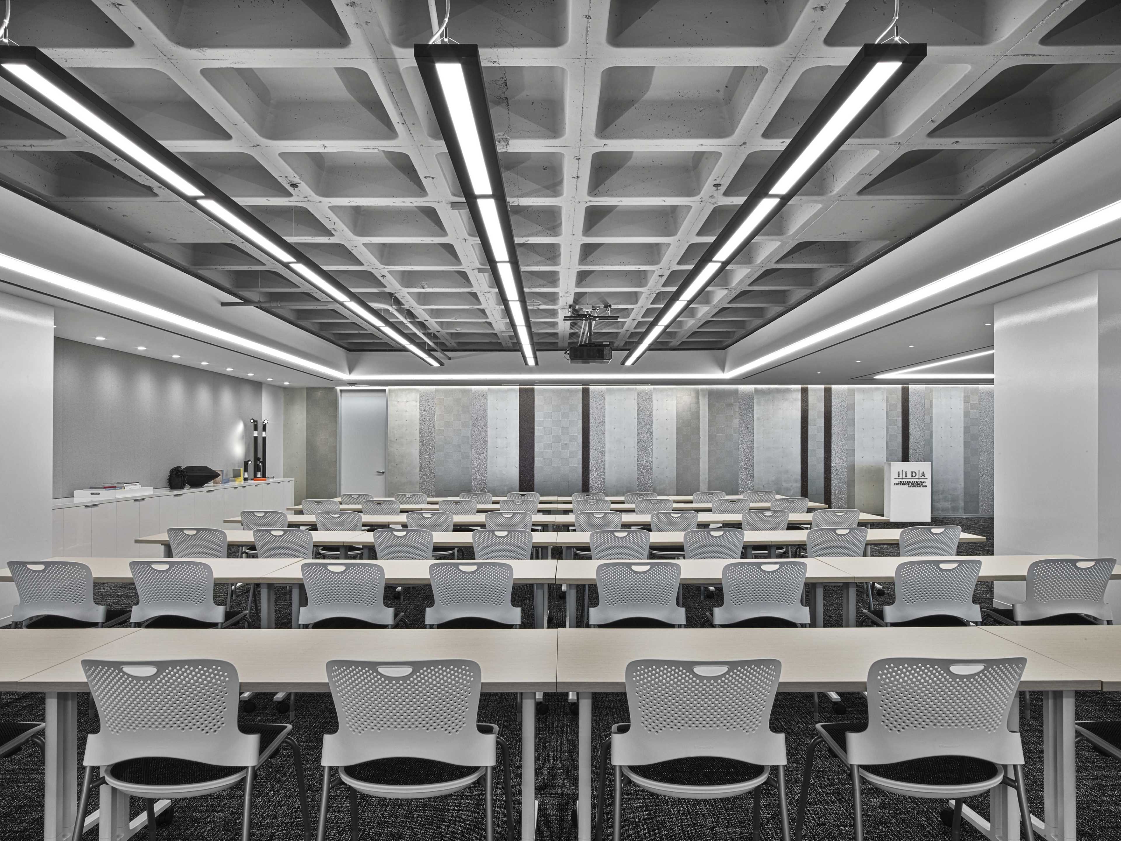 The image shows a modern conference room arranged with multiple rows of white, ergonomic chairs and long tables under a patterned ceiling with integrated lighting.