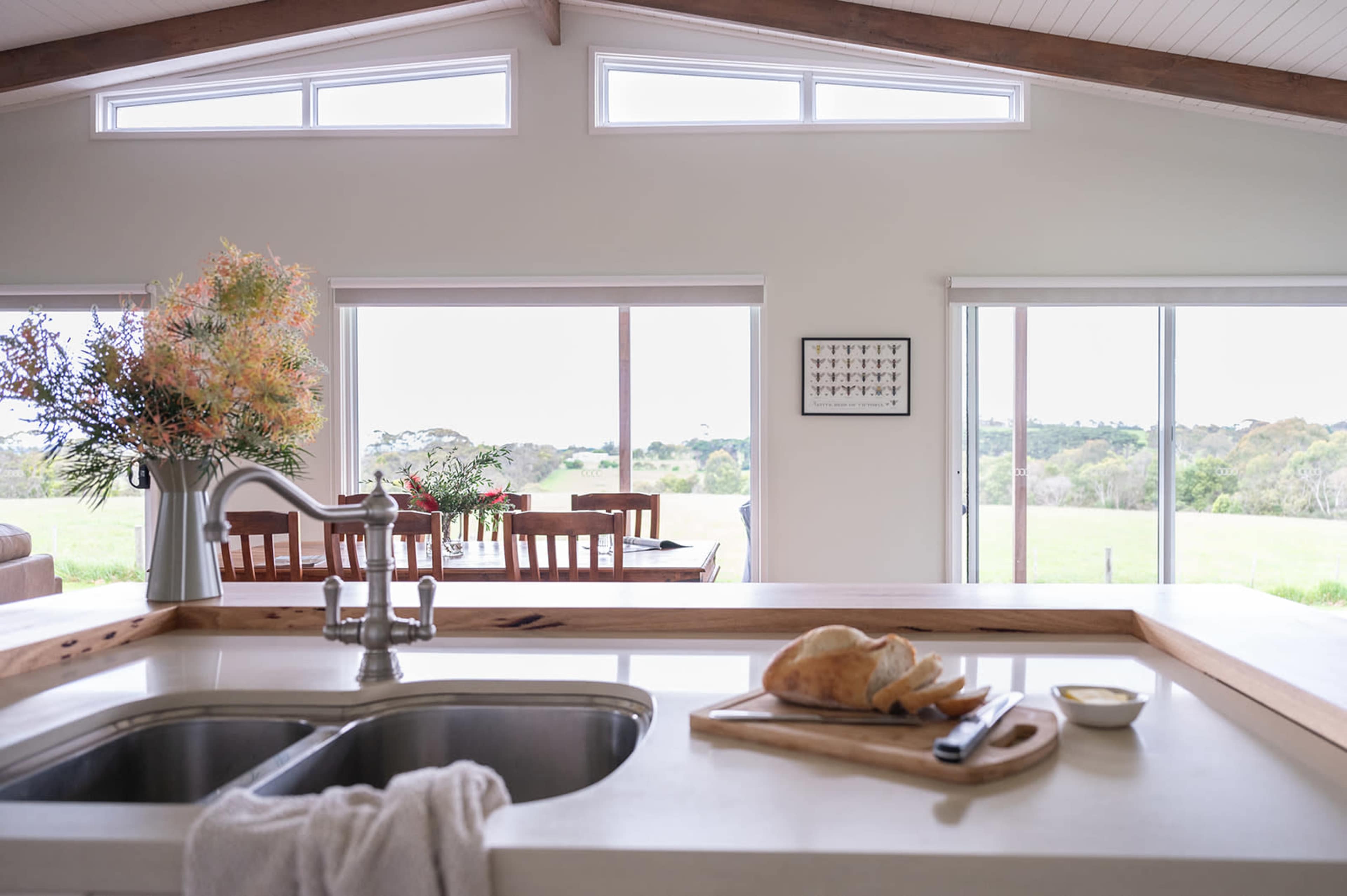 A modern kitchen features a wooden countertop with a sink, cutting board, and bread, while large windows offer a view of a green landscape.