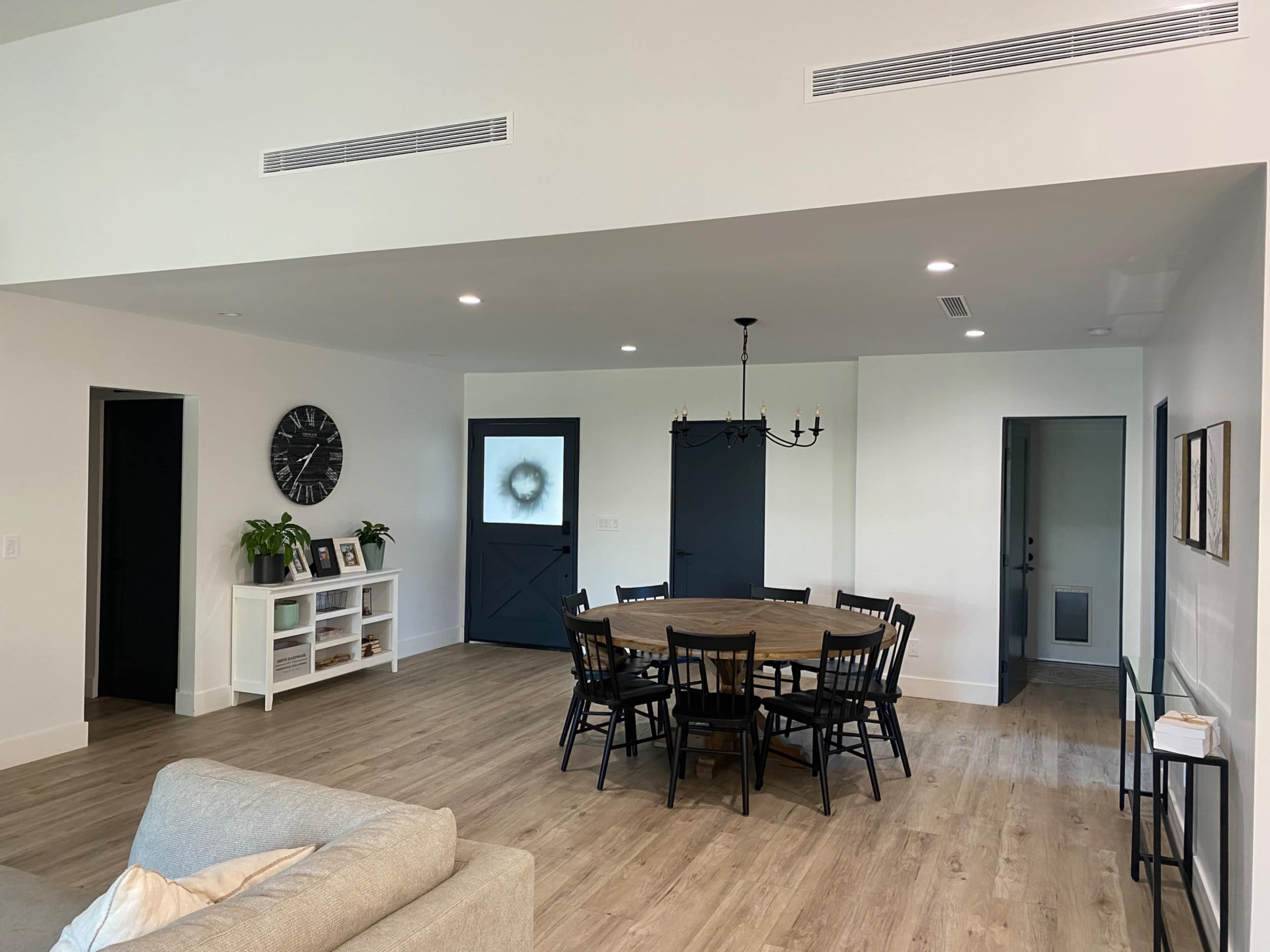 A spacious, modern living area with a round wooden dining table surrounded by black chairs, a wall clock, and a door leading to an outdoor space.