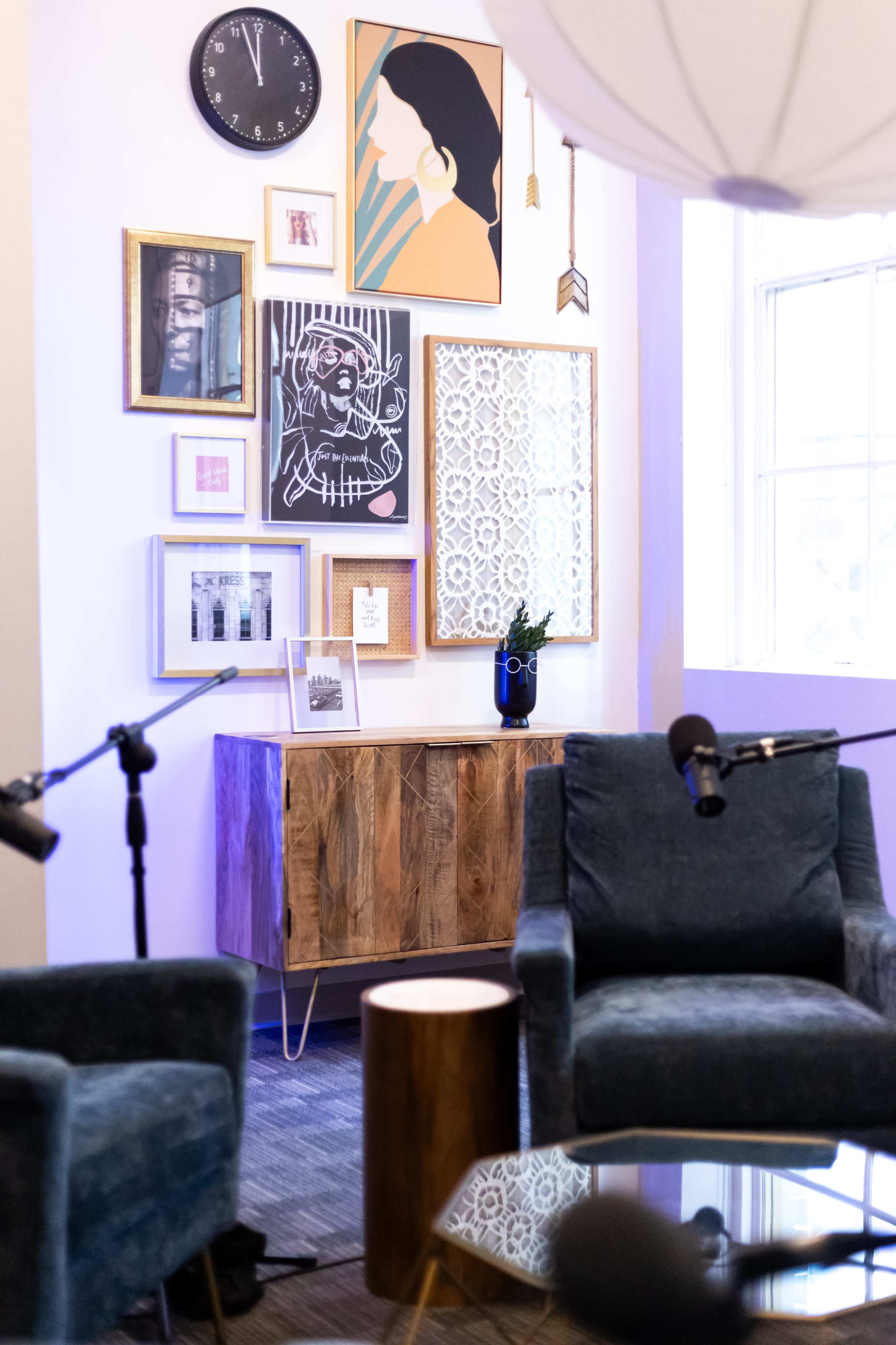 A corner of a room featuring a wooden cabinet, a variety of framed artworks on the wall, a clock, a plant, and two upholstered chairs with a small table in between.