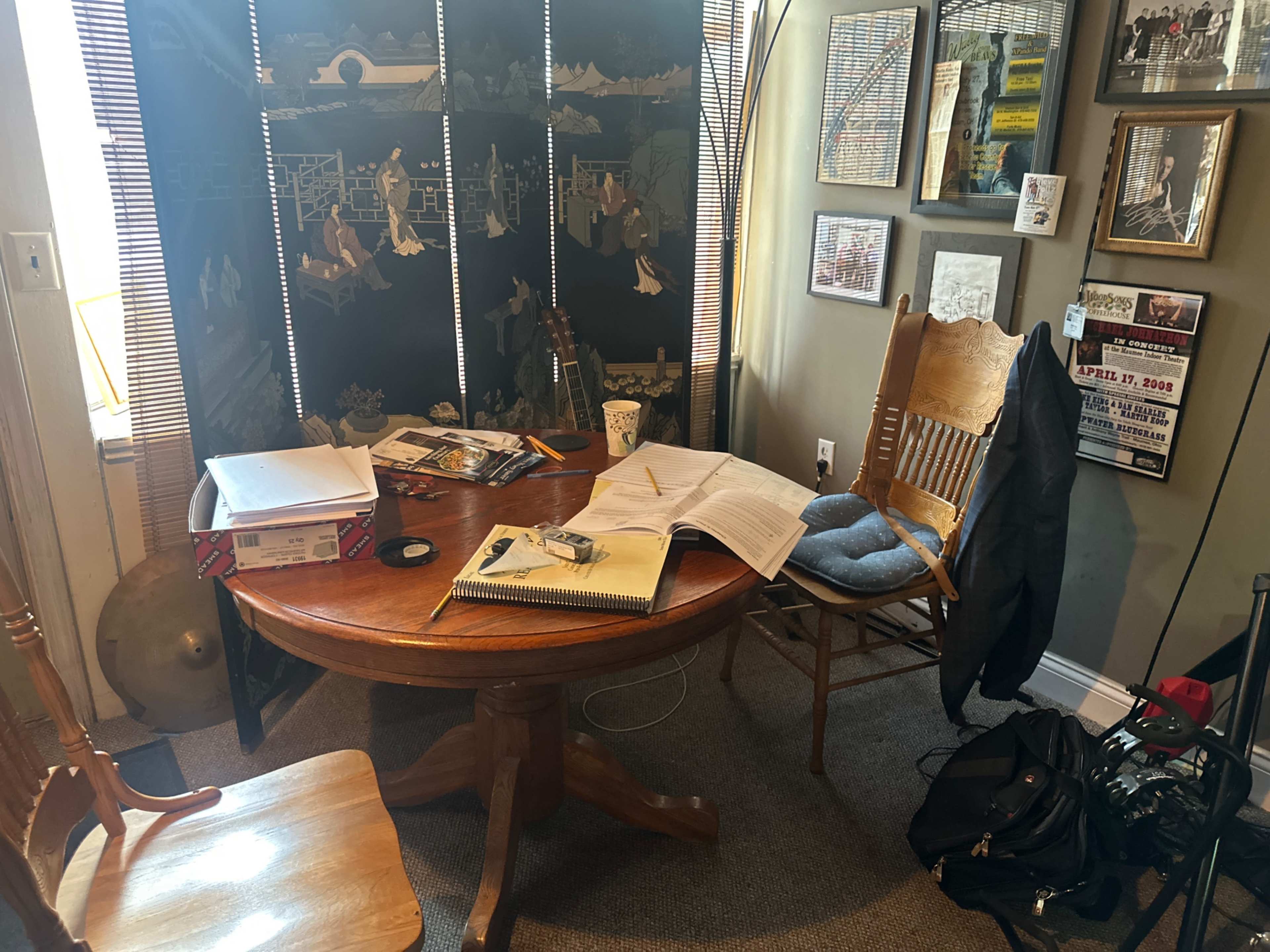 The image shows a cluttered wooden table with an open book, some papers, a coffee cup, and a chair beside it, all set in a small room adorned with framed pictures and a folding screen.