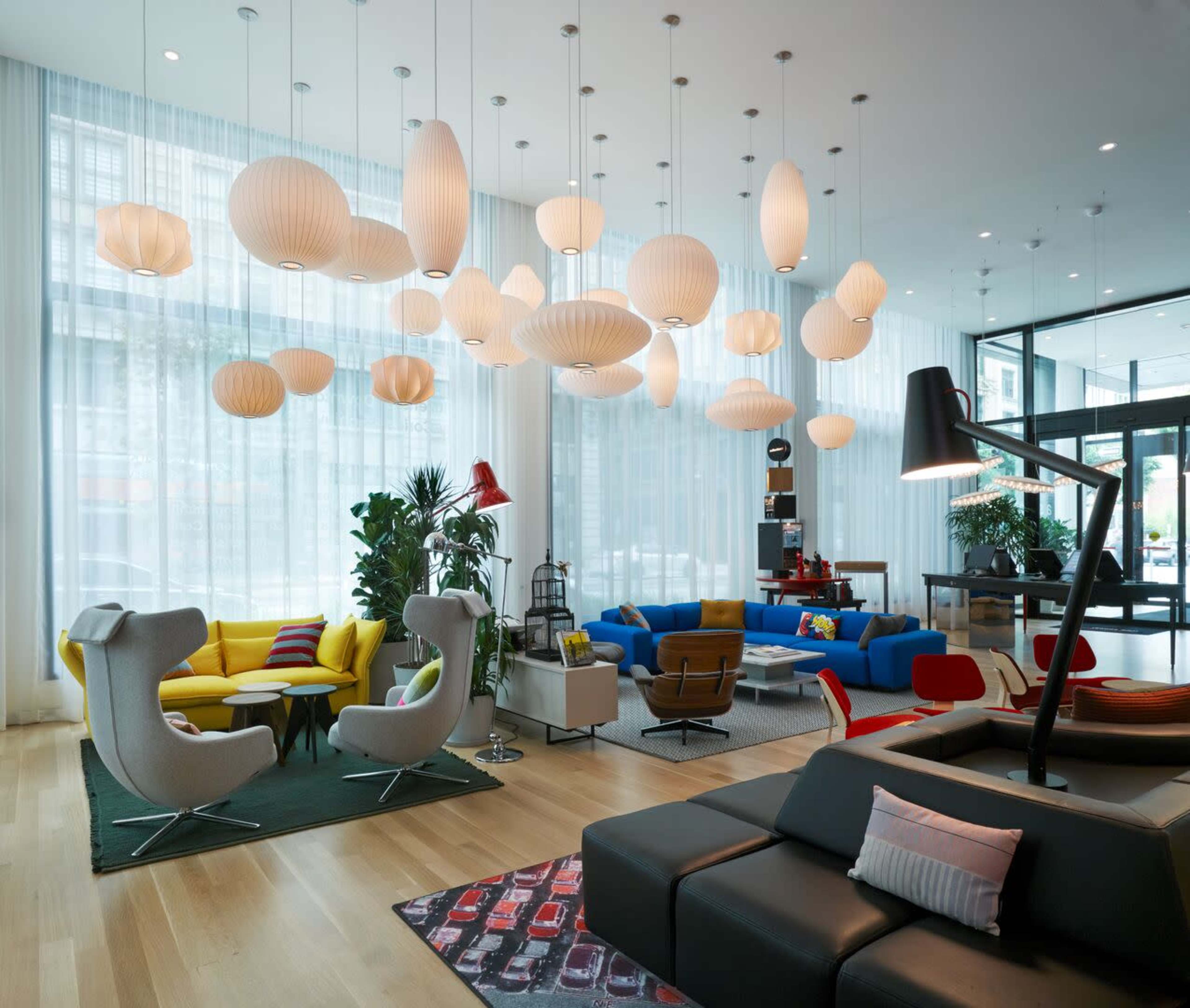 The image depicts a modern, spacious lounge area with various seating arrangements featuring colorful furniture and a display of pendant lights overhead.