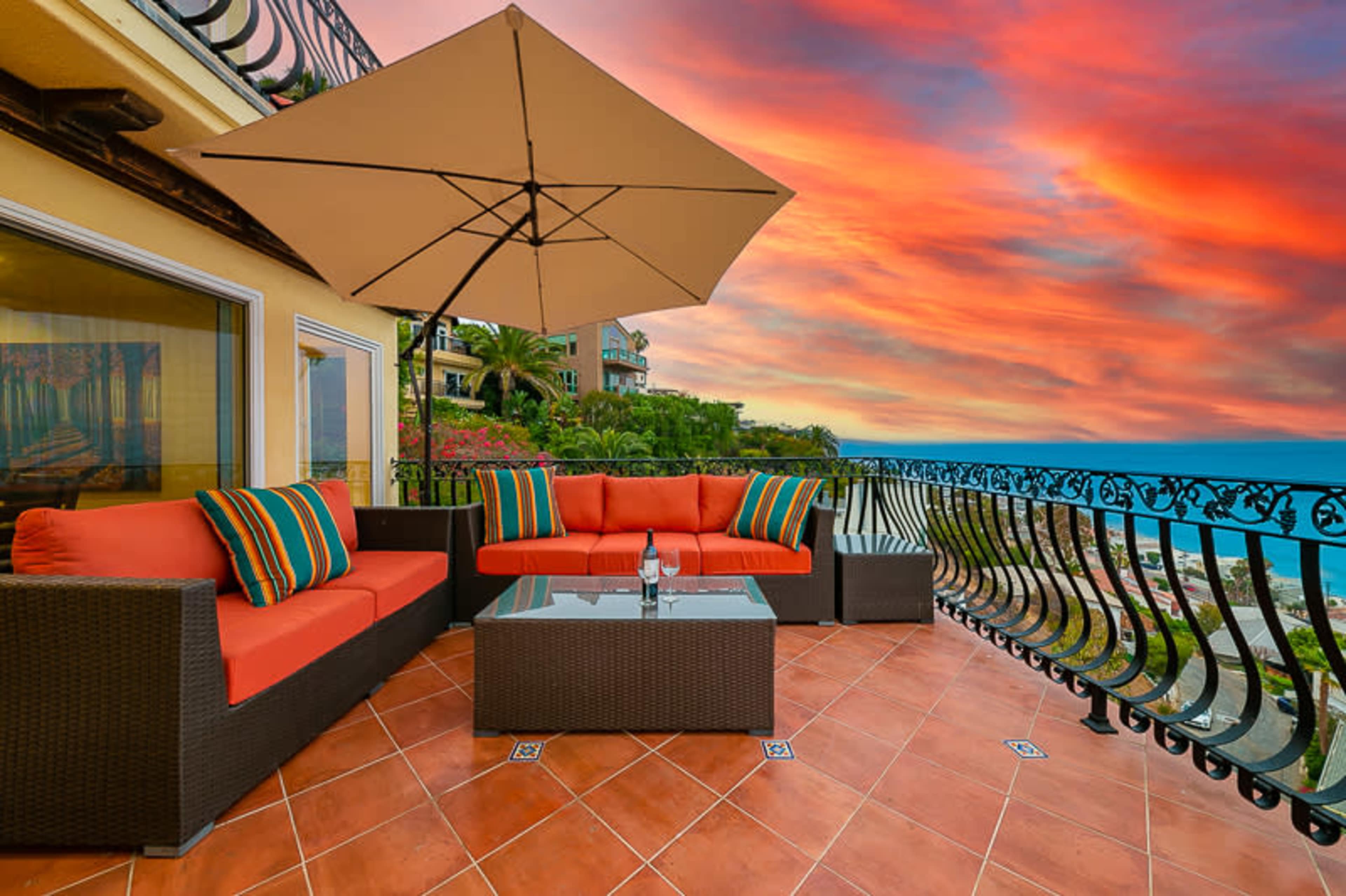 A patio with orange seating and a glass table is set against a backdrop of a vibrant sunset over the ocean.