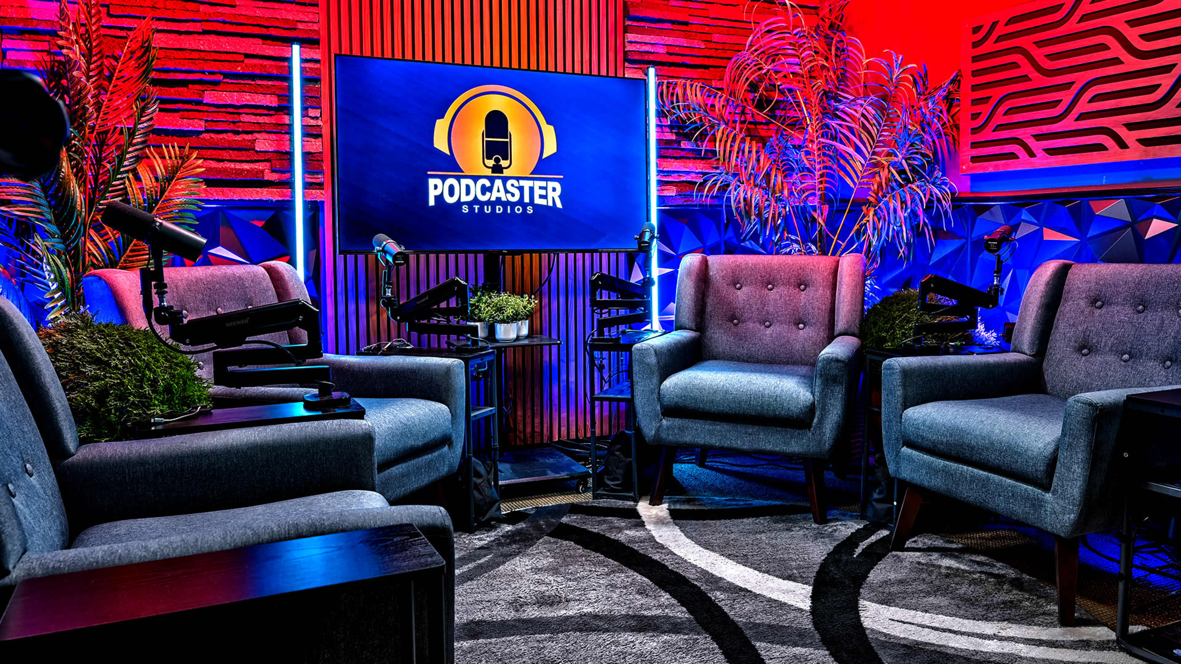 The image shows a podcast studio featuring four grey armchairs arranged around a central area with a patterned rug, a large screen displaying the "Podcaster Studios" logo, and decorative plants in a brightly lit environment with red and blue hues.