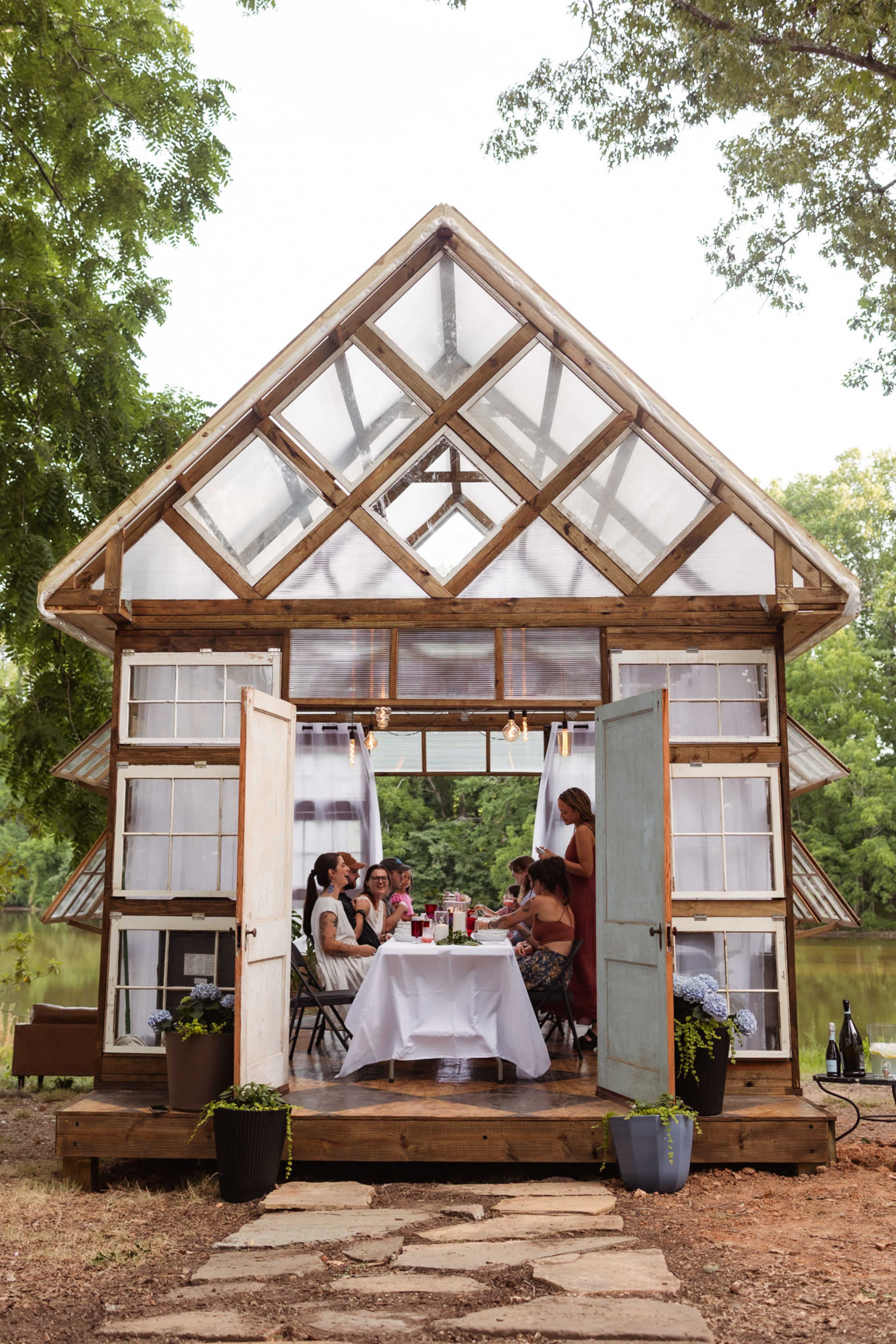 A group of six people sit around a table set for dinner inside a glass-enclosed house by a river.