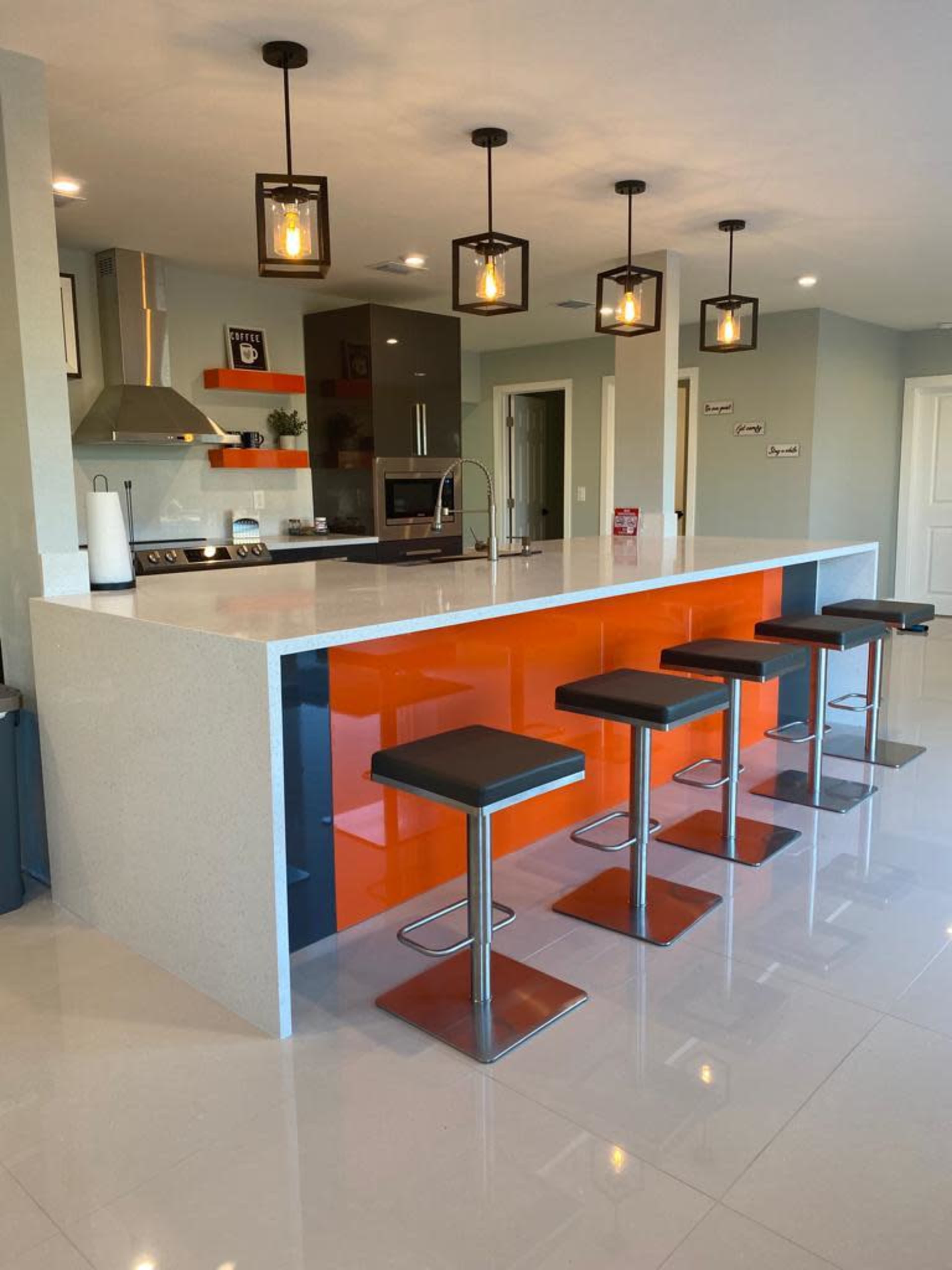 A modern kitchen features a central island with an orange accent and five black bar stools arranged around it.