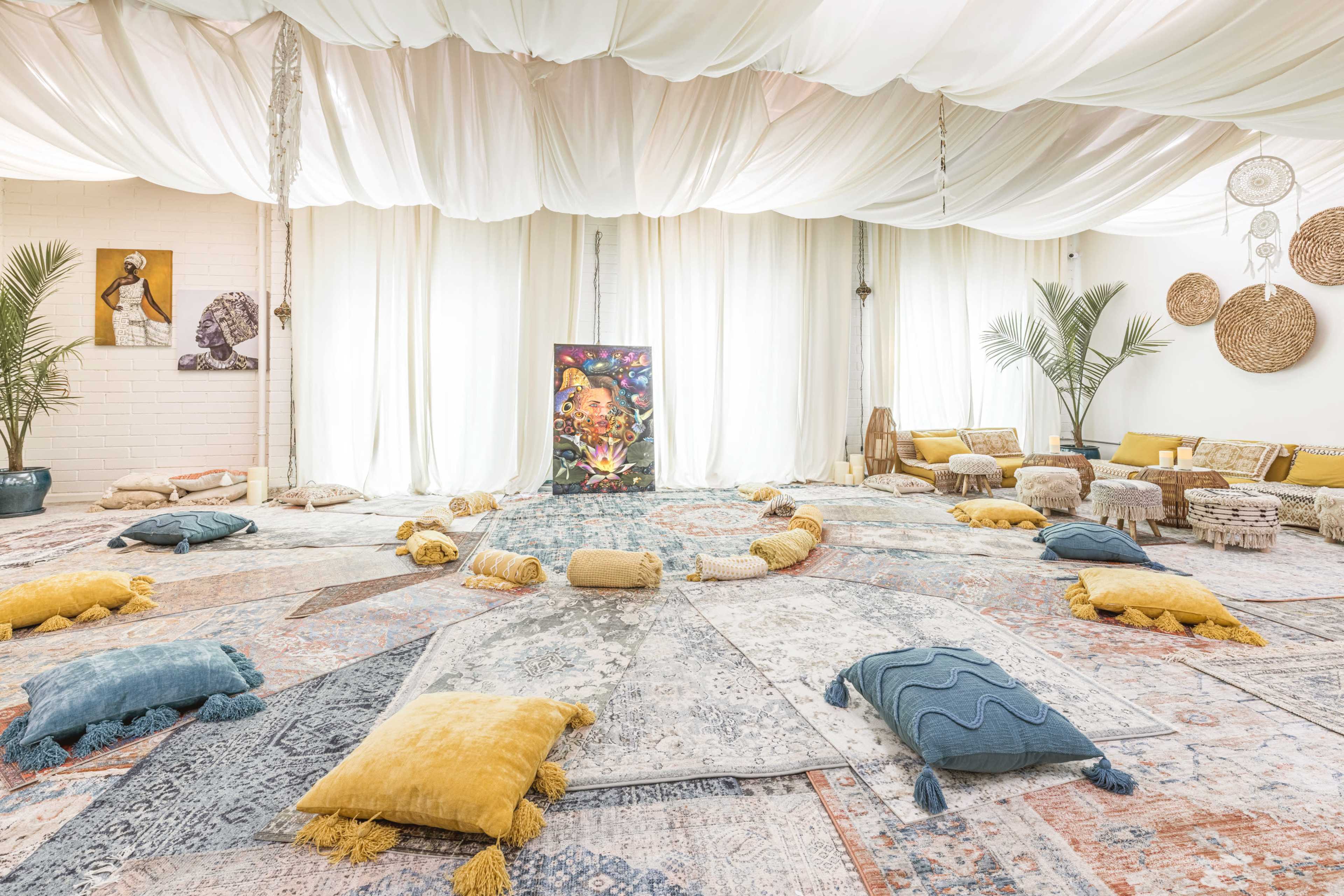 A spacious indoor area decorated with rugs, cushions, and a central artwork, surrounded by plants and soft drapery.