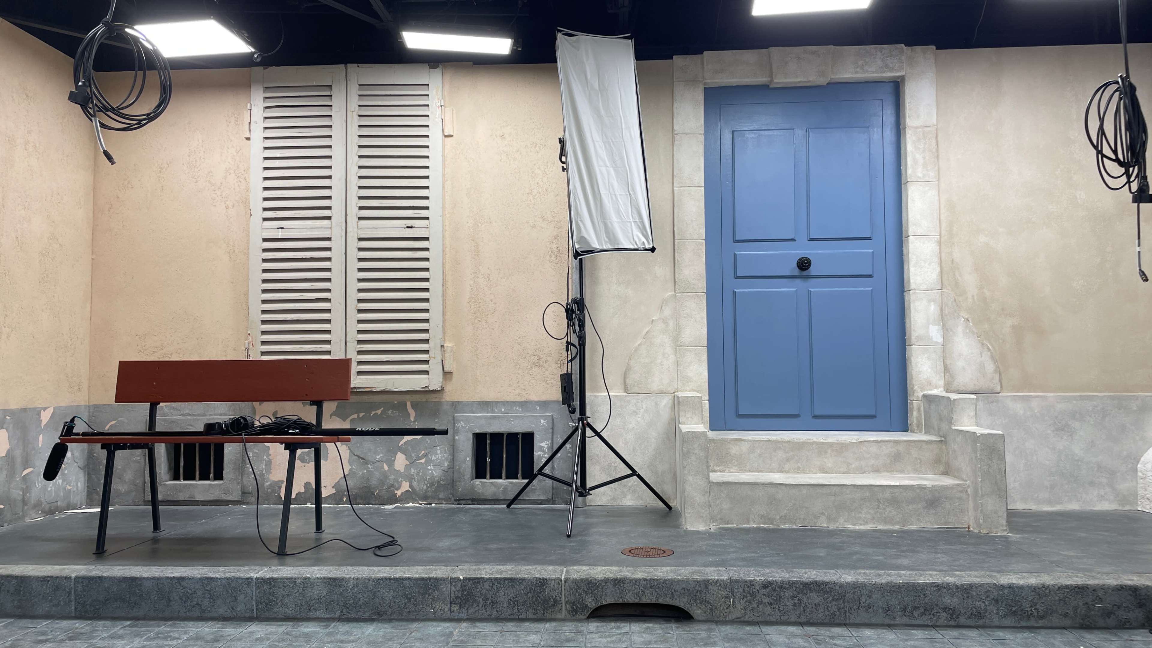 A staged set with a blue door, a wooden bench, a tall backdrop with curtains, and lighting equipment, arranged on a textured floor.
