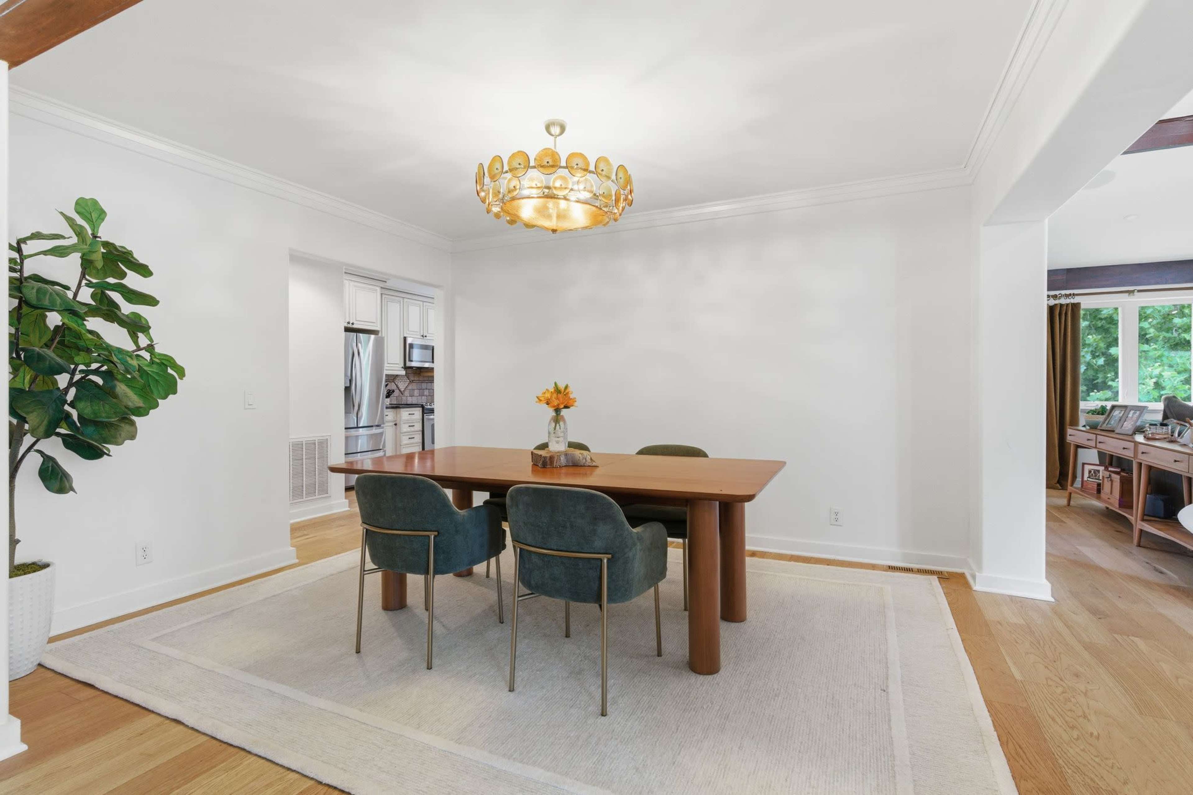 A modern dining room features a wooden table surrounded by green chairs, complemented by a large chandelier and a potted plant.