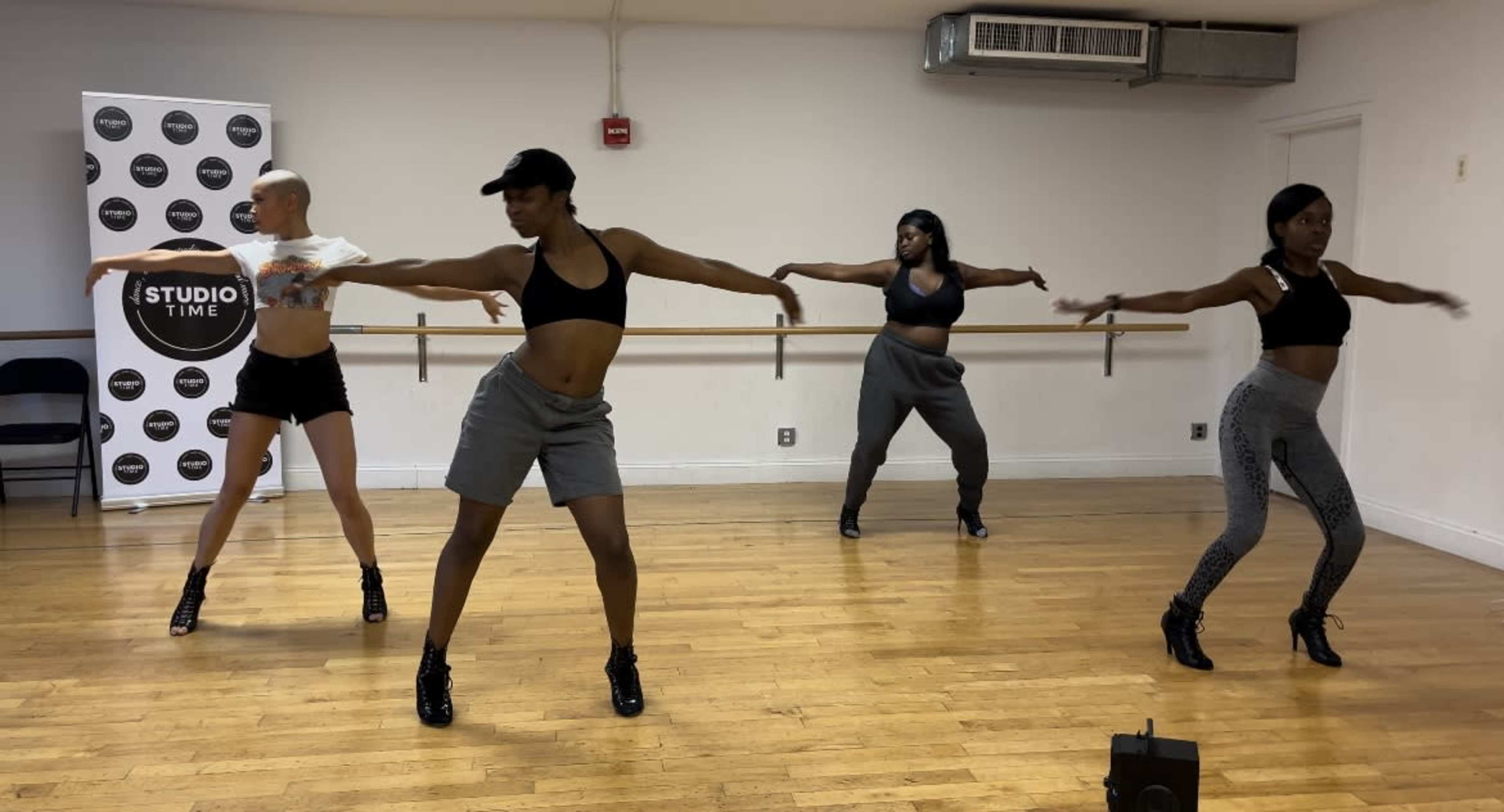 A group of four dancers performs a routine in a dance studio with wooden floors and a mirrored wall.