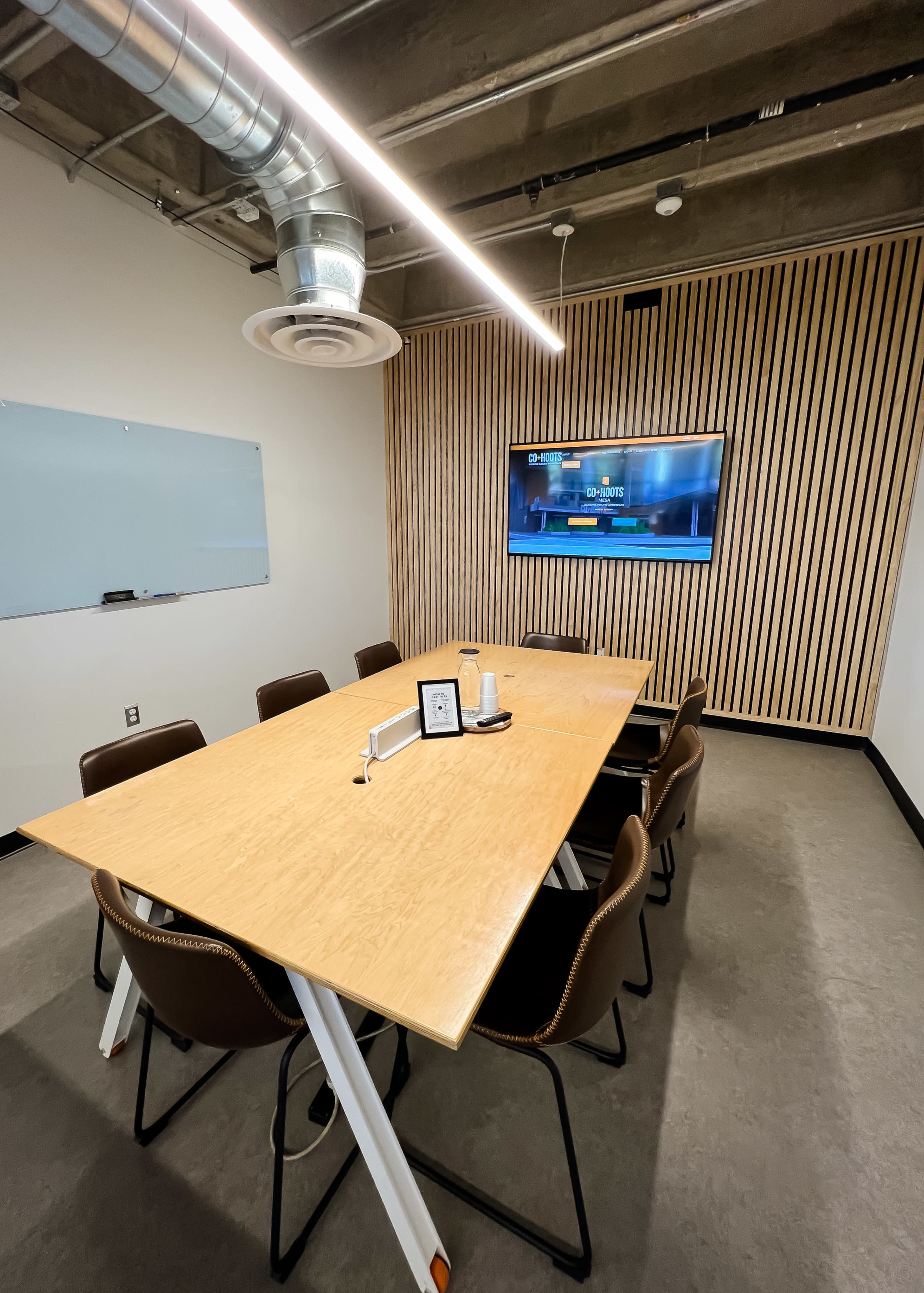 A modern conference room features a long wooden table with chairs, a wall-mounted TV, a whiteboard, and wooden paneling.