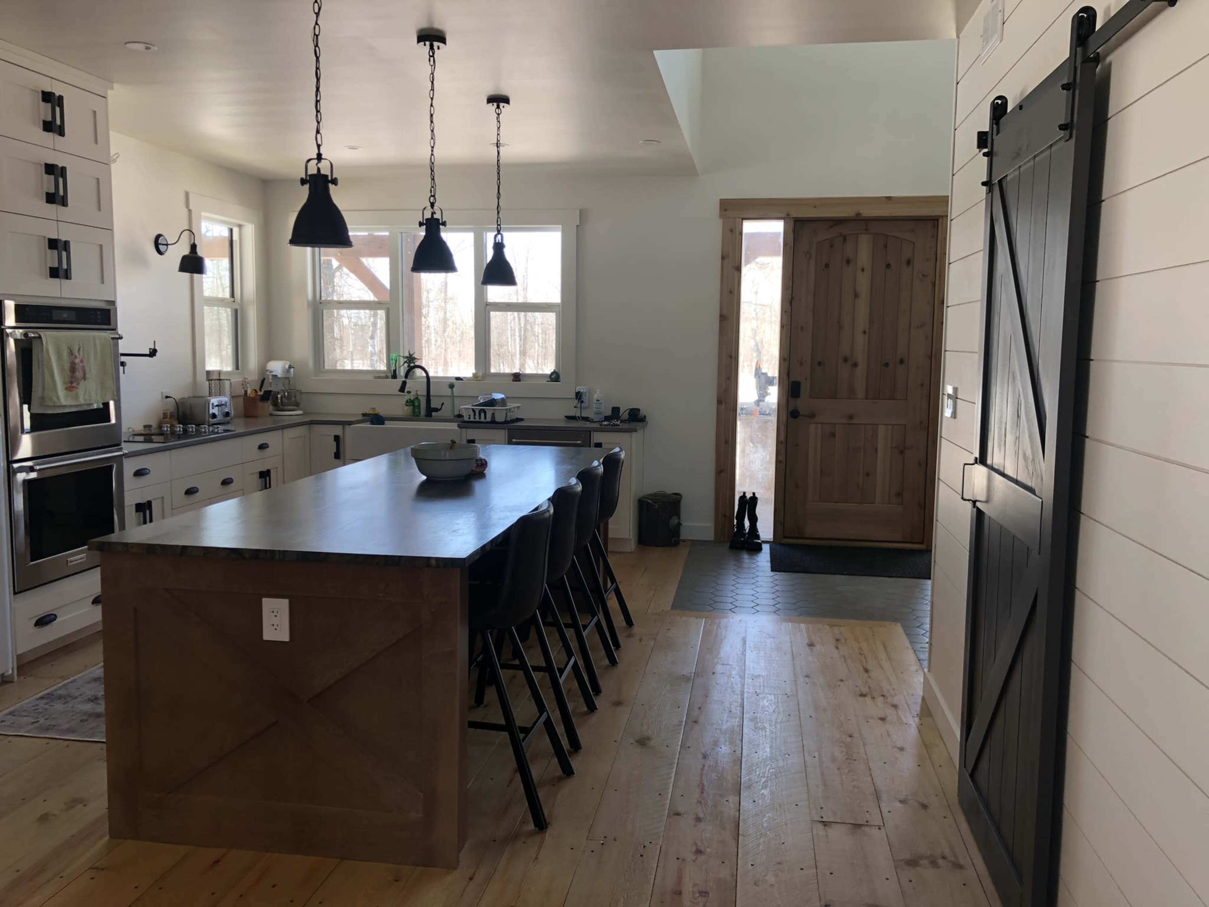 A modern kitchen with a central island, black pendant lights, and a wooden door leading to an entryway.