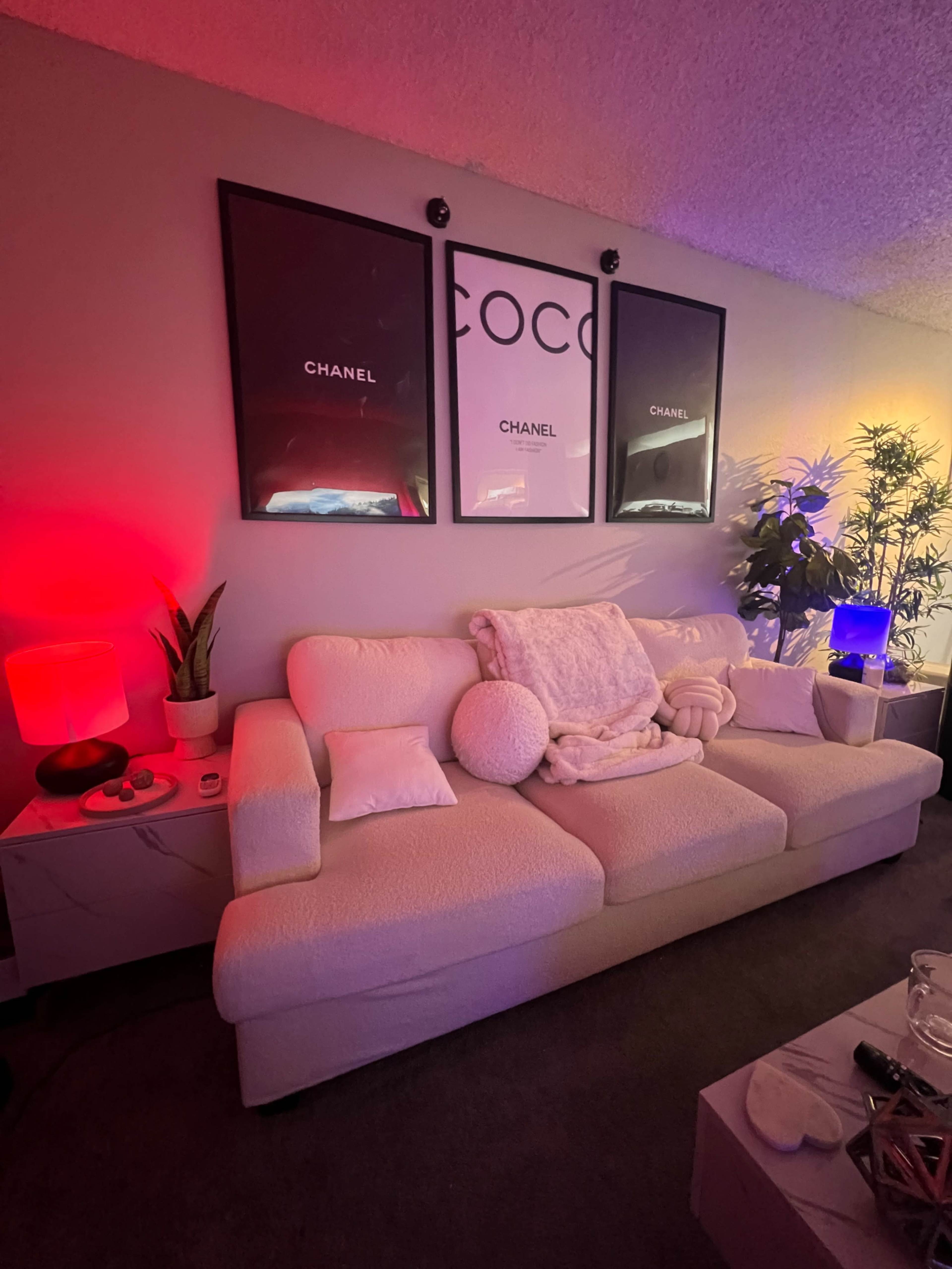 A cozy living room with a light-colored couch adorned with a throw blanket and decorative pillows, flanked by framed Chanel posters and illuminated by colorful lamps.