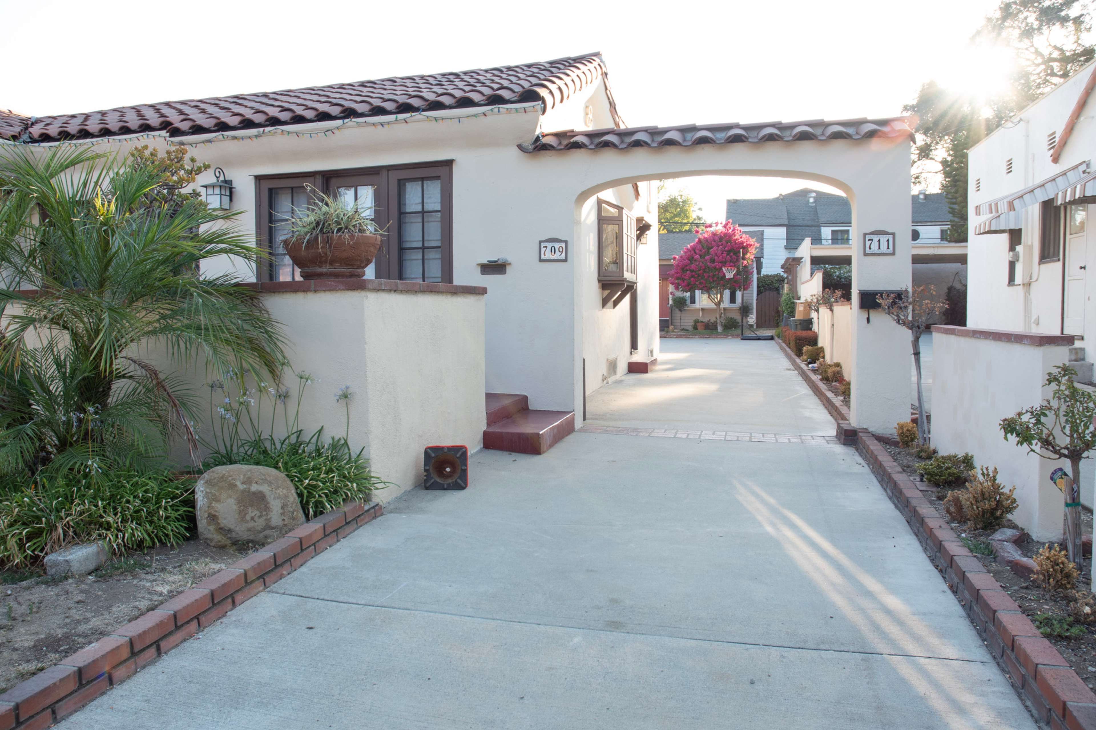 A quiet residential street with houses on either side, lined by a paved driveway leading to two mailbox posts.