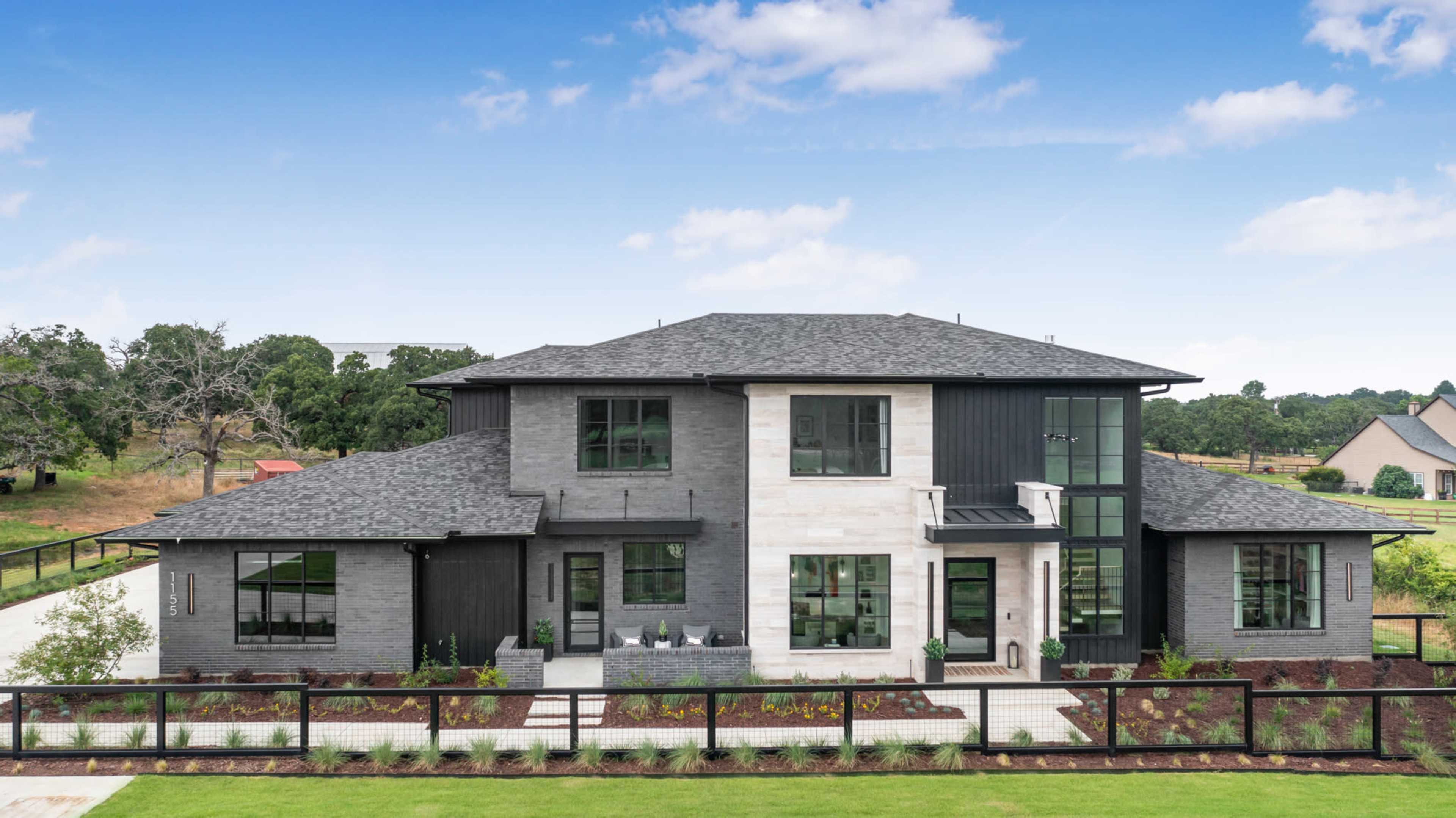 A modern two-story house featuring a mix of dark brick and light stone exteriors, surrounded by landscaped grounds and a wooden fence.