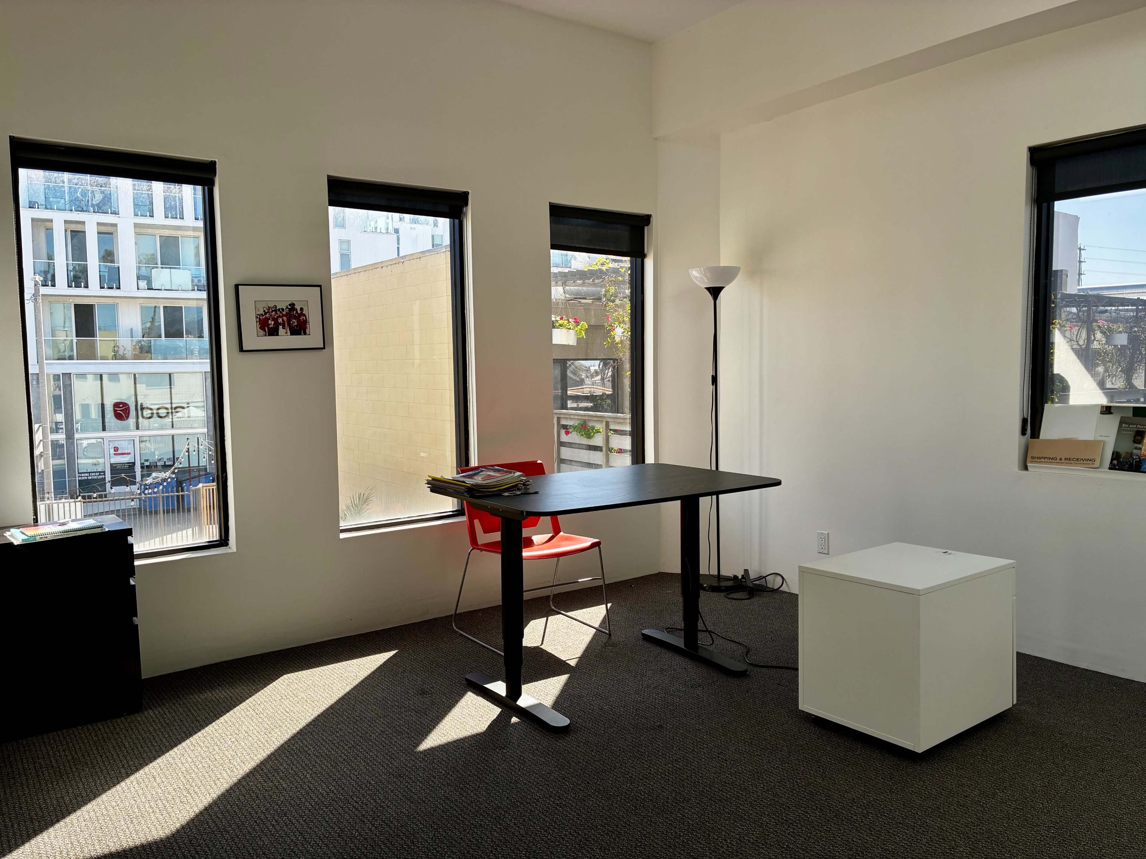 The image shows a minimalist office space with a black table, a red chair, and natural light streaming in through three windows.
