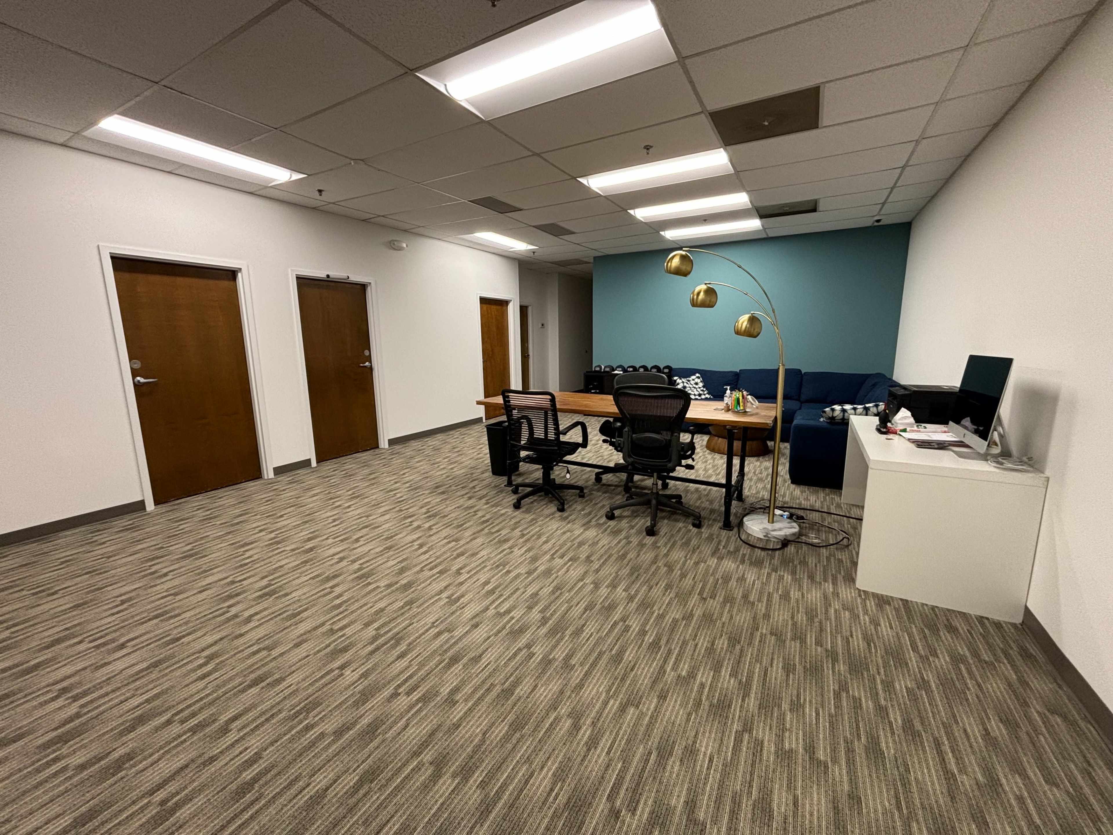 A modern office space featuring two closed doors, a desk with two chairs, a decorative floor lamp, and a couch against a teal wall.