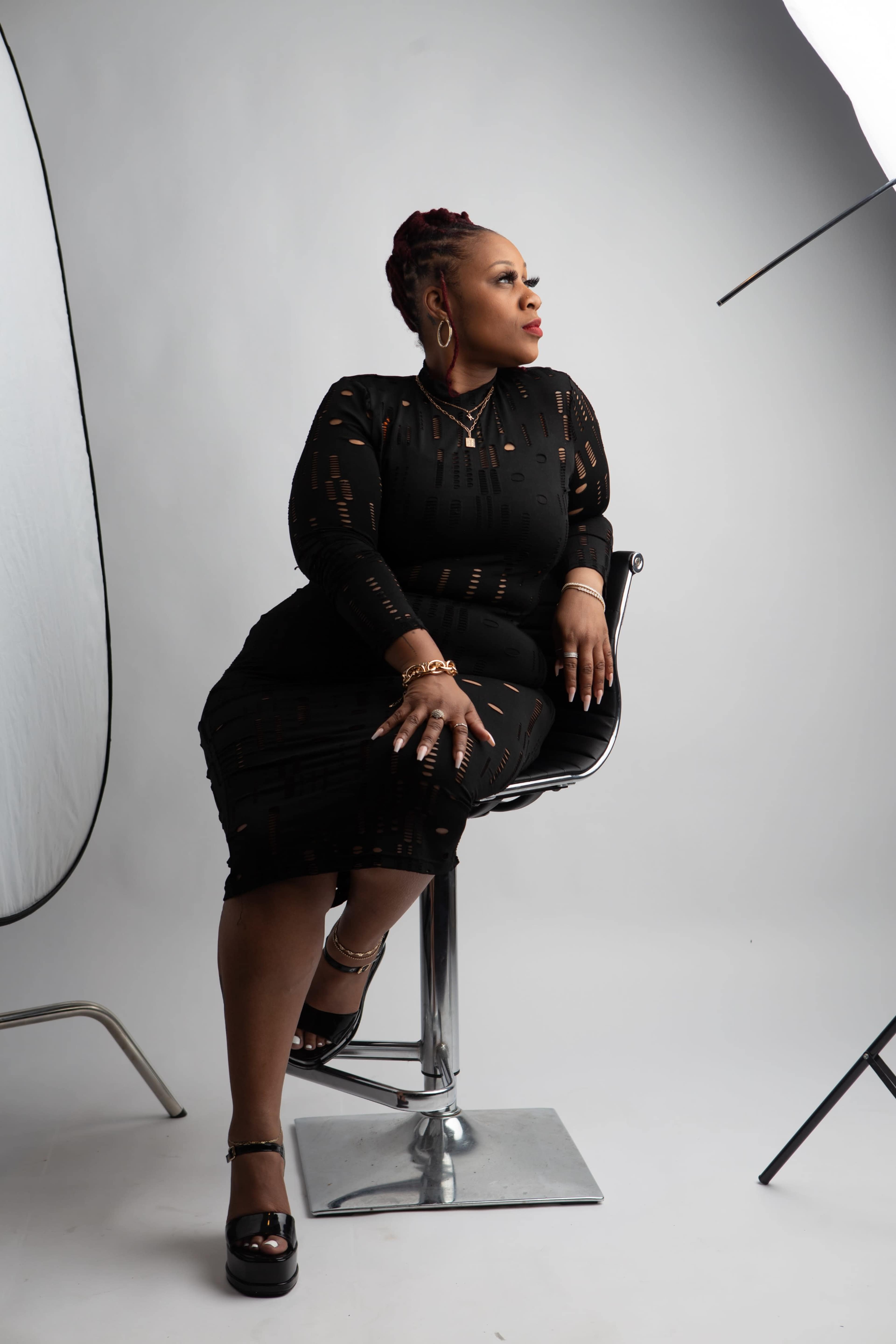 A woman in a black dress sits elegantly on a chrome bar stool, facing to the side while illuminated by studio lighting.