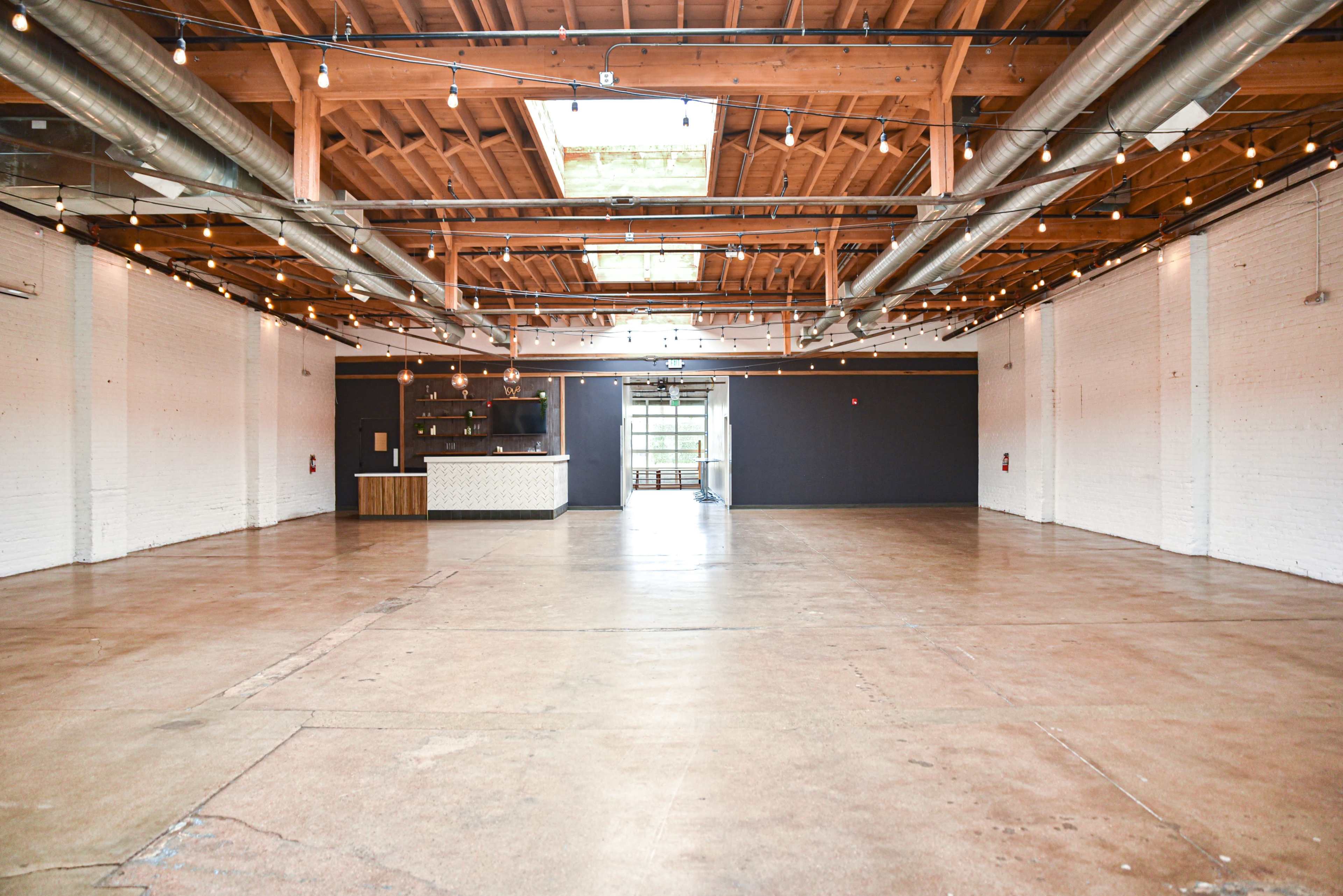 The image shows a spacious, vacant interior with polished concrete flooring, exposed wooden beams, and industrial-style lighting fixtures.