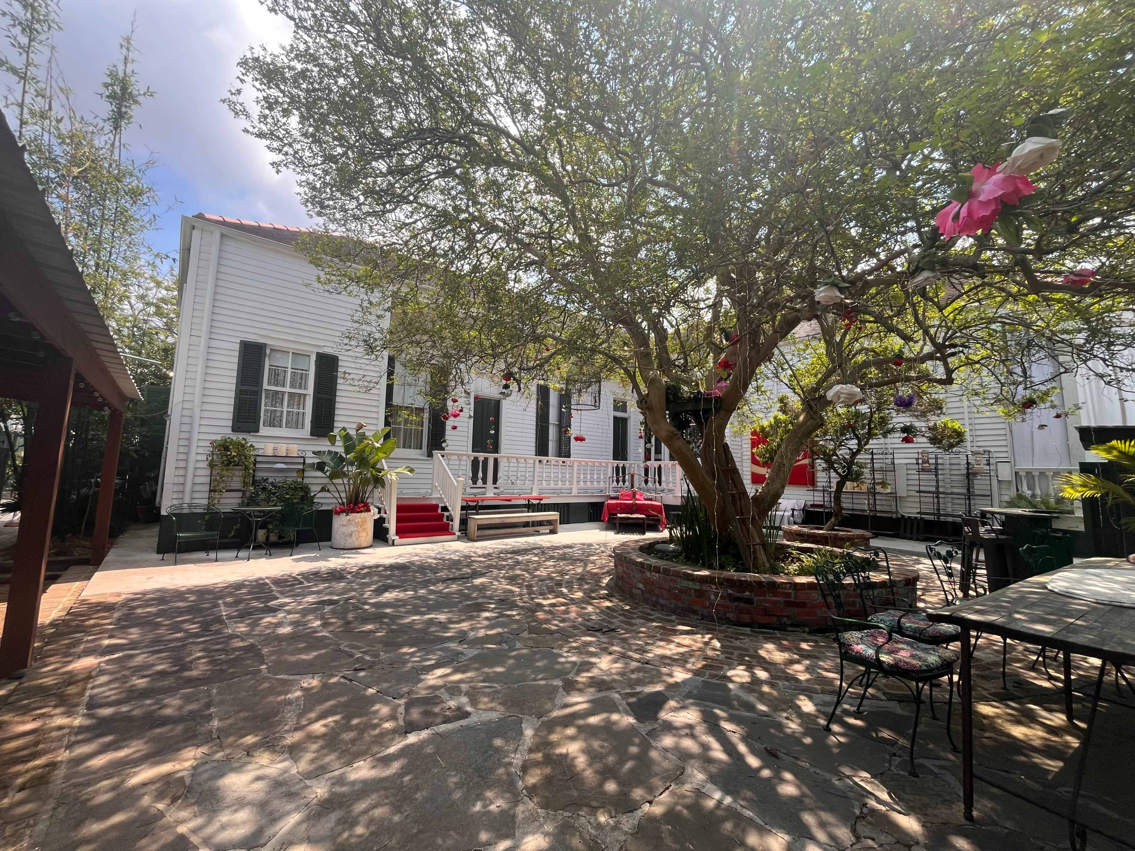 The image shows a courtyard with a brick planter and a large tree, surrounding a two-story white house with green shutters and adorned with decorations.