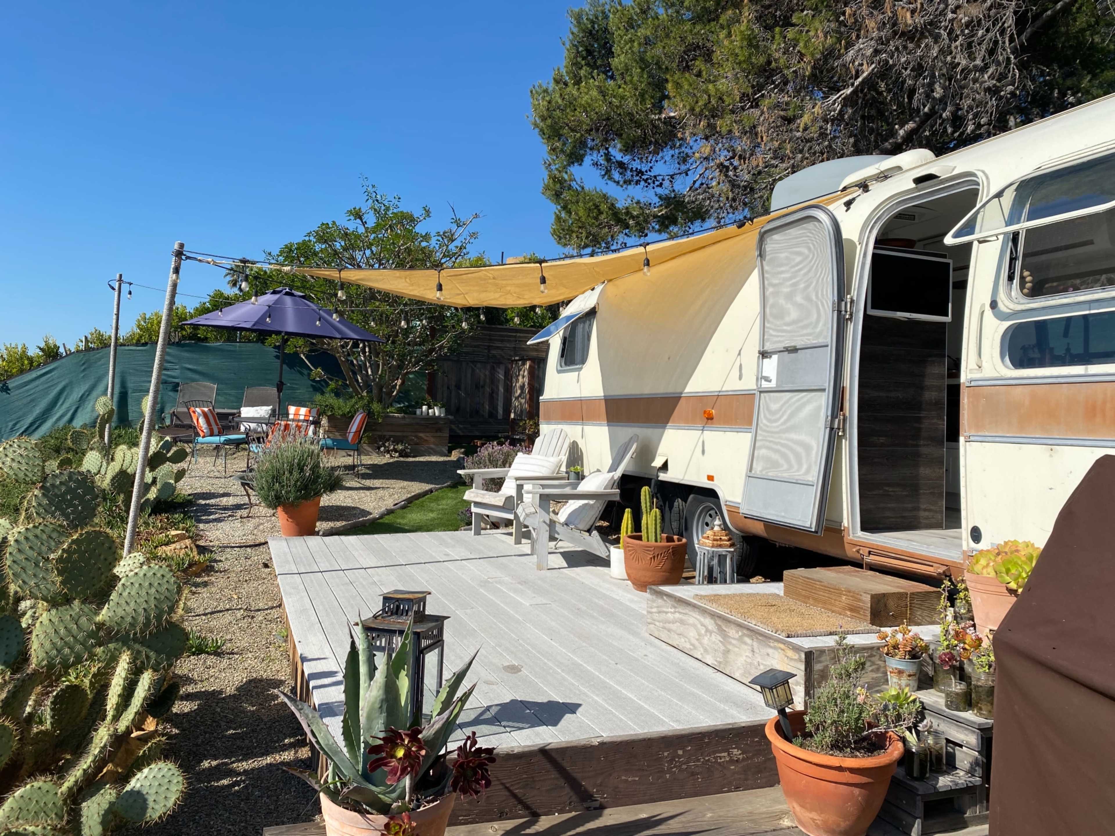 A vintage trailer is parked next to a wooden deck adorned with potted plants, a table and chairs under an umbrella, and surrounded by shrubs and trees.