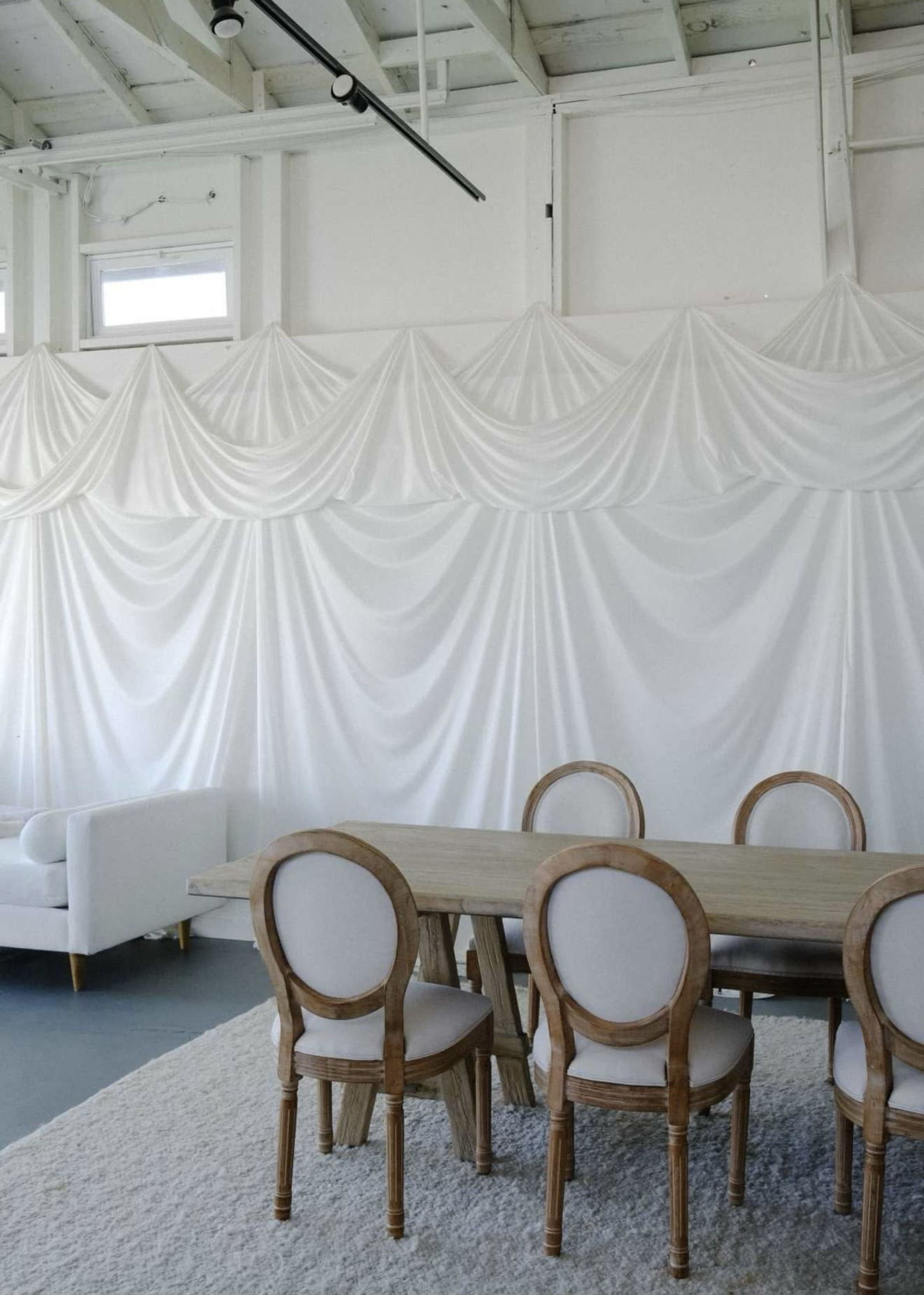 A dining area features a wooden table surrounded by upholstered chairs, set against a backdrop of draped white fabric and a light-colored carpet.