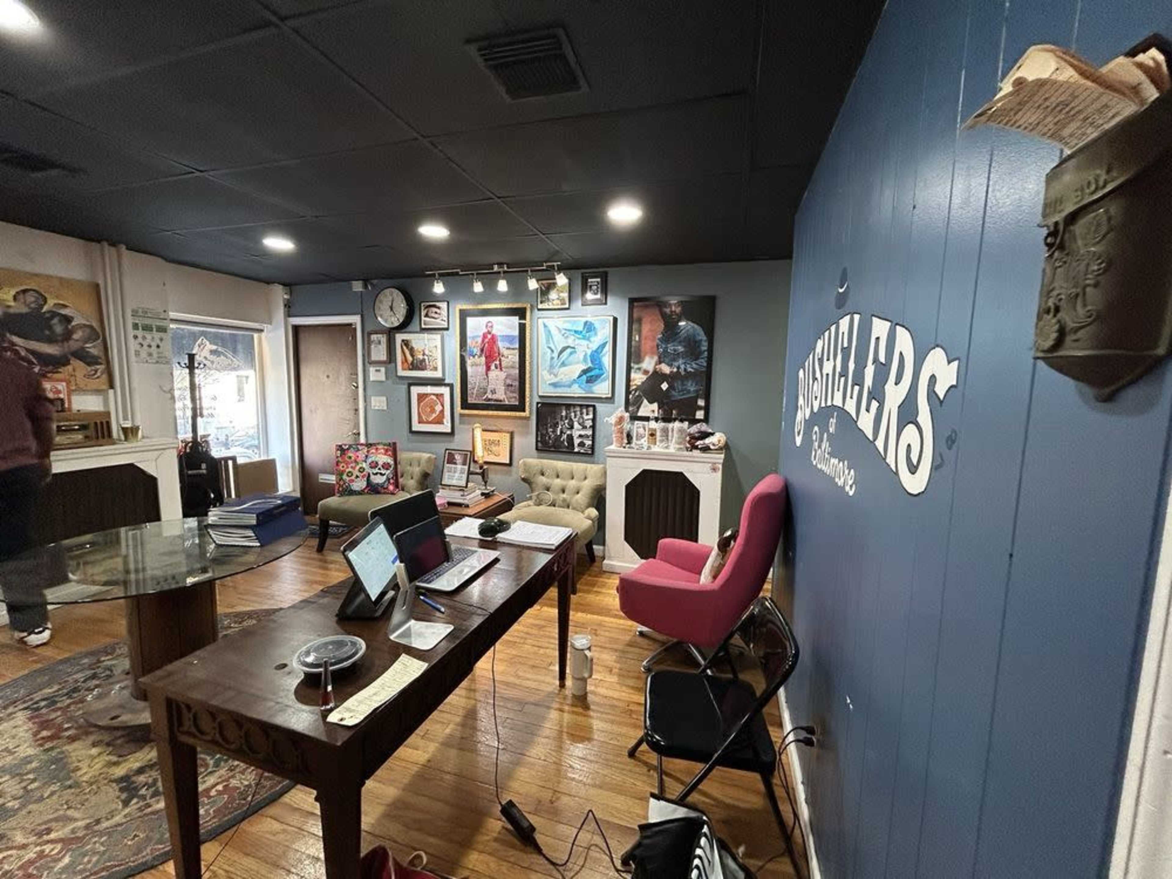 The image shows an office space with a large table, various artworks on the walls, a pink chair, and wooden flooring.