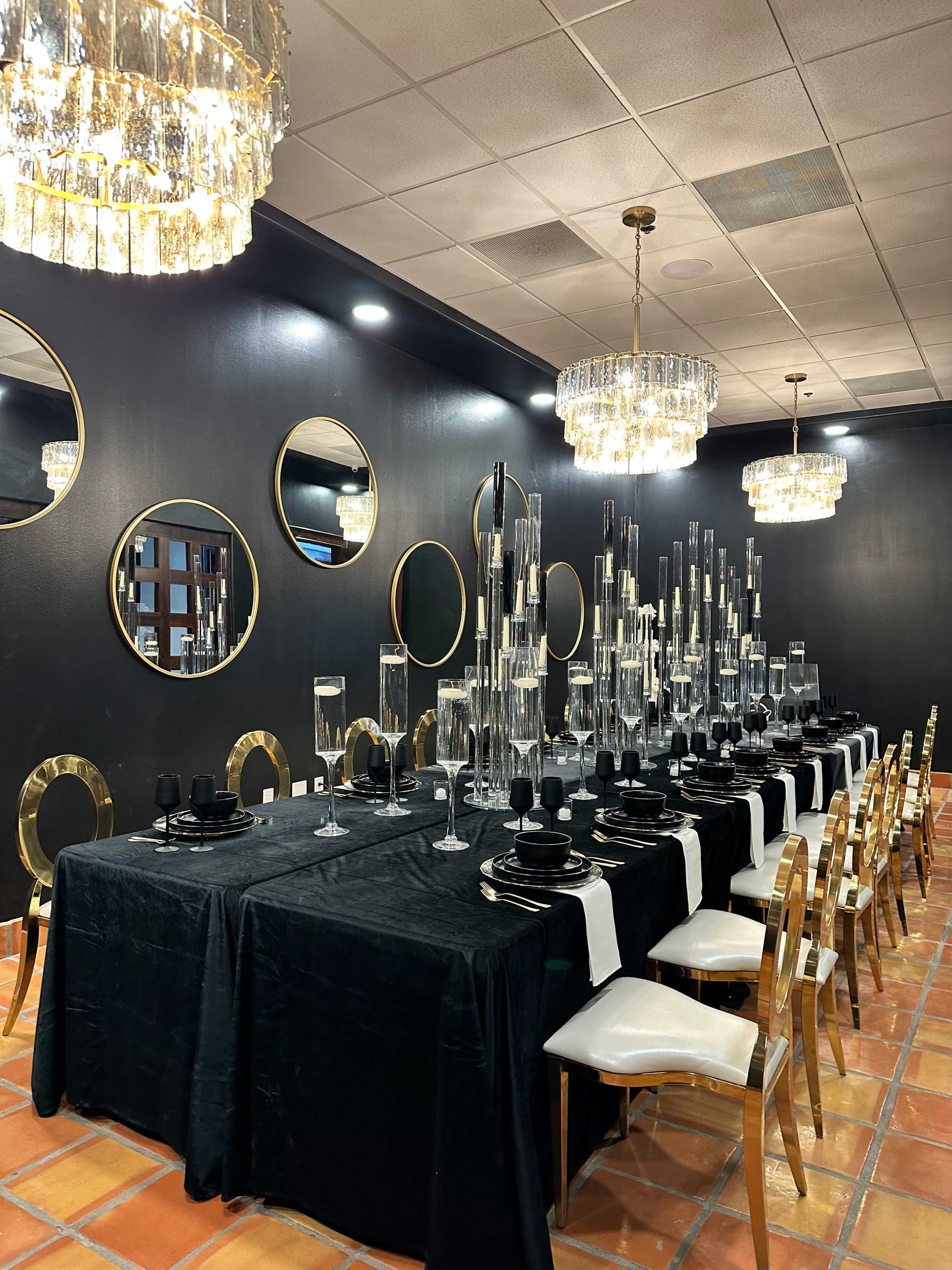 A long table is set for dining, adorned with black tablecloths, elegant glassware, and candle holders, in a room featuring black walls and multiple round mirrors.
