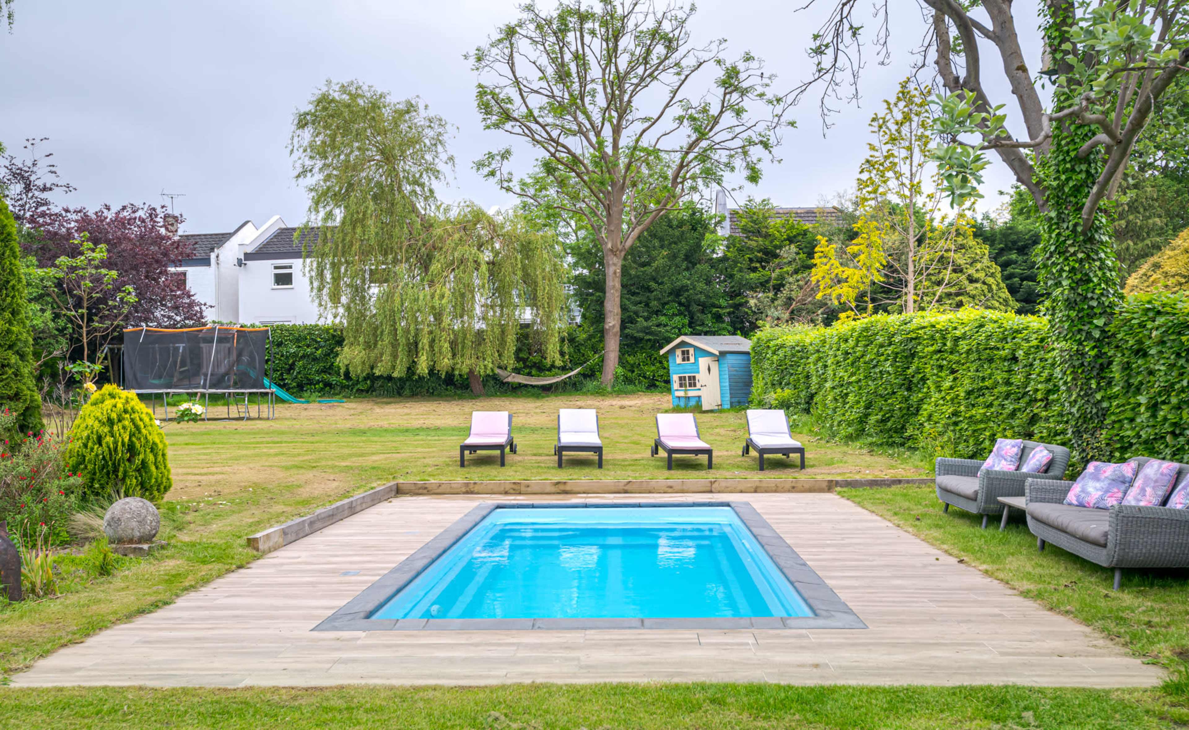 A backyard featuring a rectangular swimming pool surrounded by wooden decking, with loungers arranged nearby and a play set in the background.
