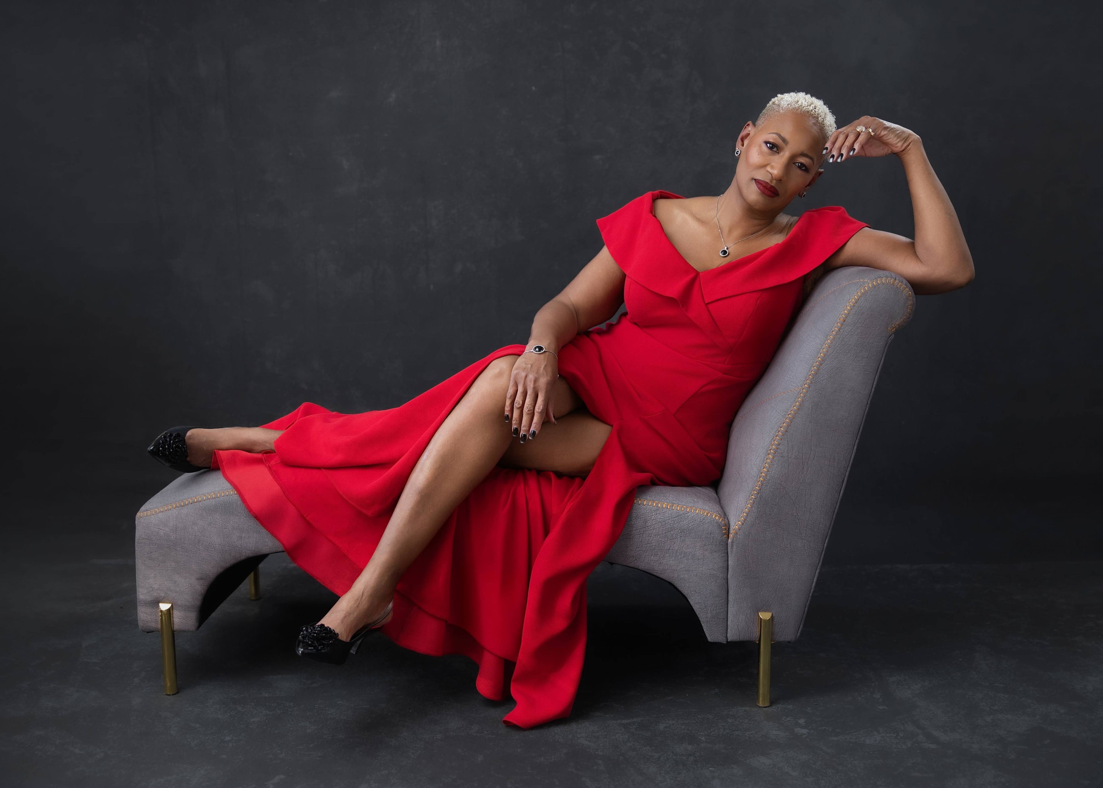 A woman in a red dress reclines on a grey upholstered chaise lounge against a dark backdrop.