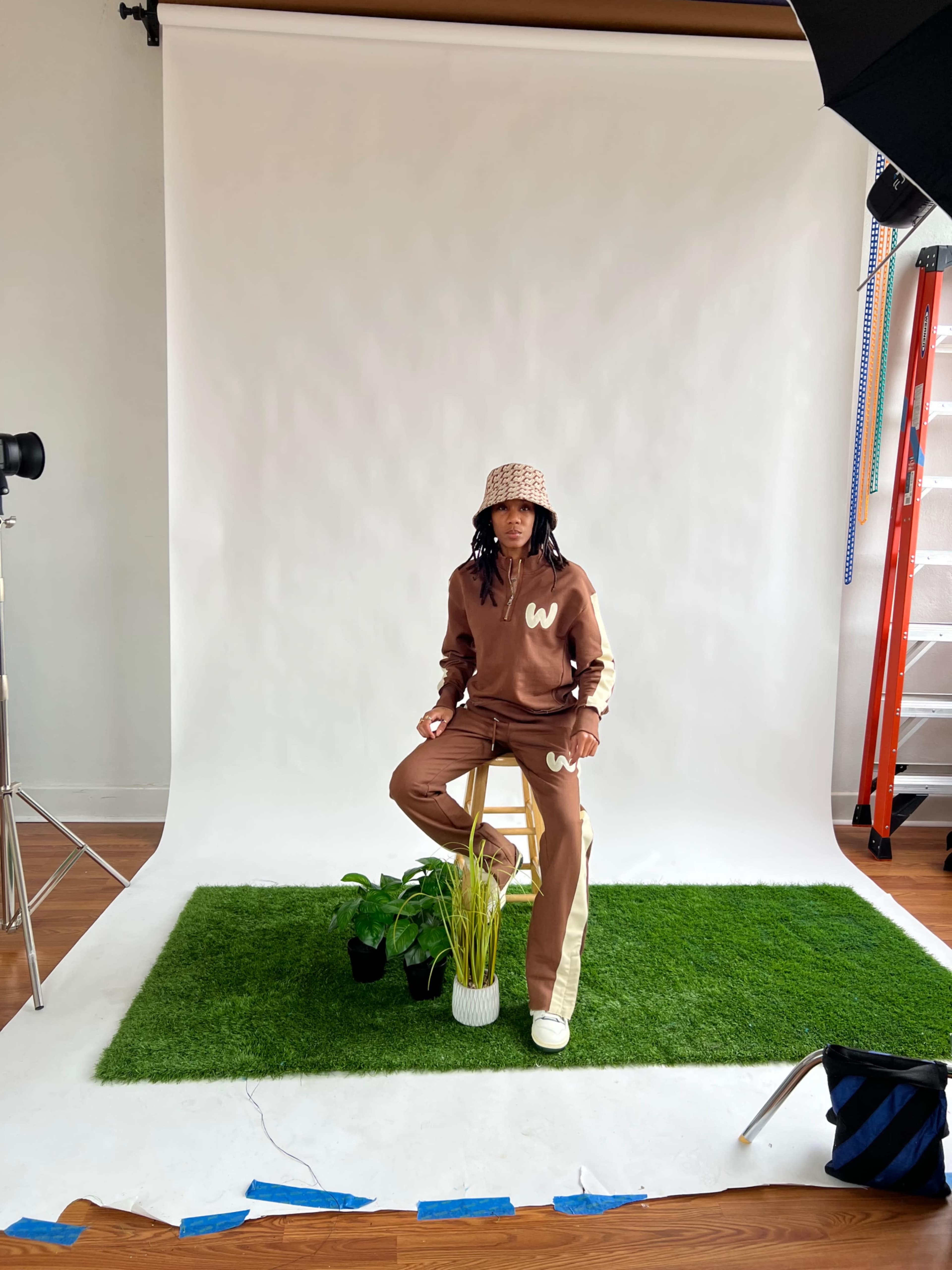 A person sits on a stool in a studio setting with a green grass-like backdrop, wearing a brown outfit and a checkered hat, surrounded by potted plants.