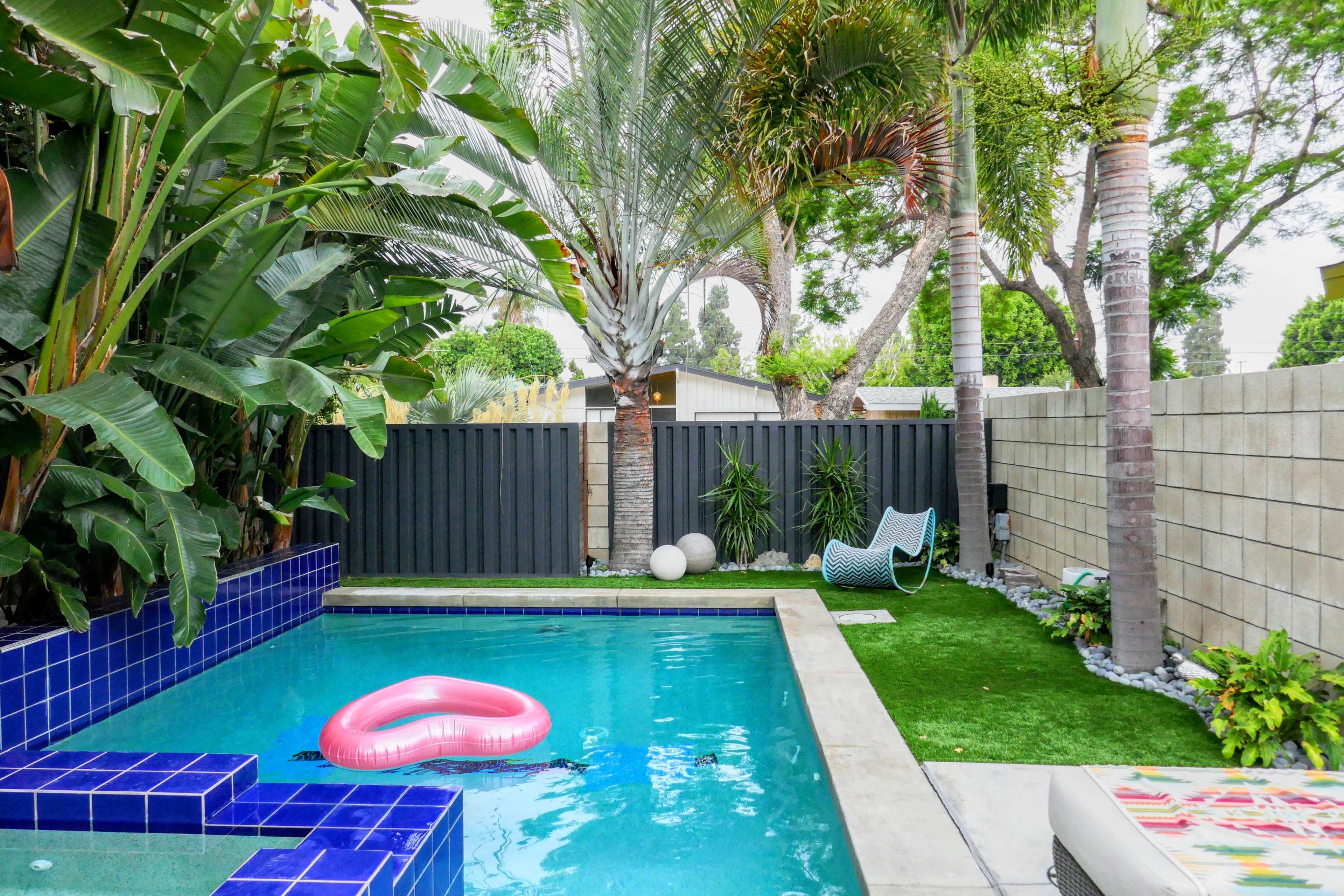 A swimming pool with a pink inflatable float is surrounded by tropical plants and palm trees in a backyard setting.