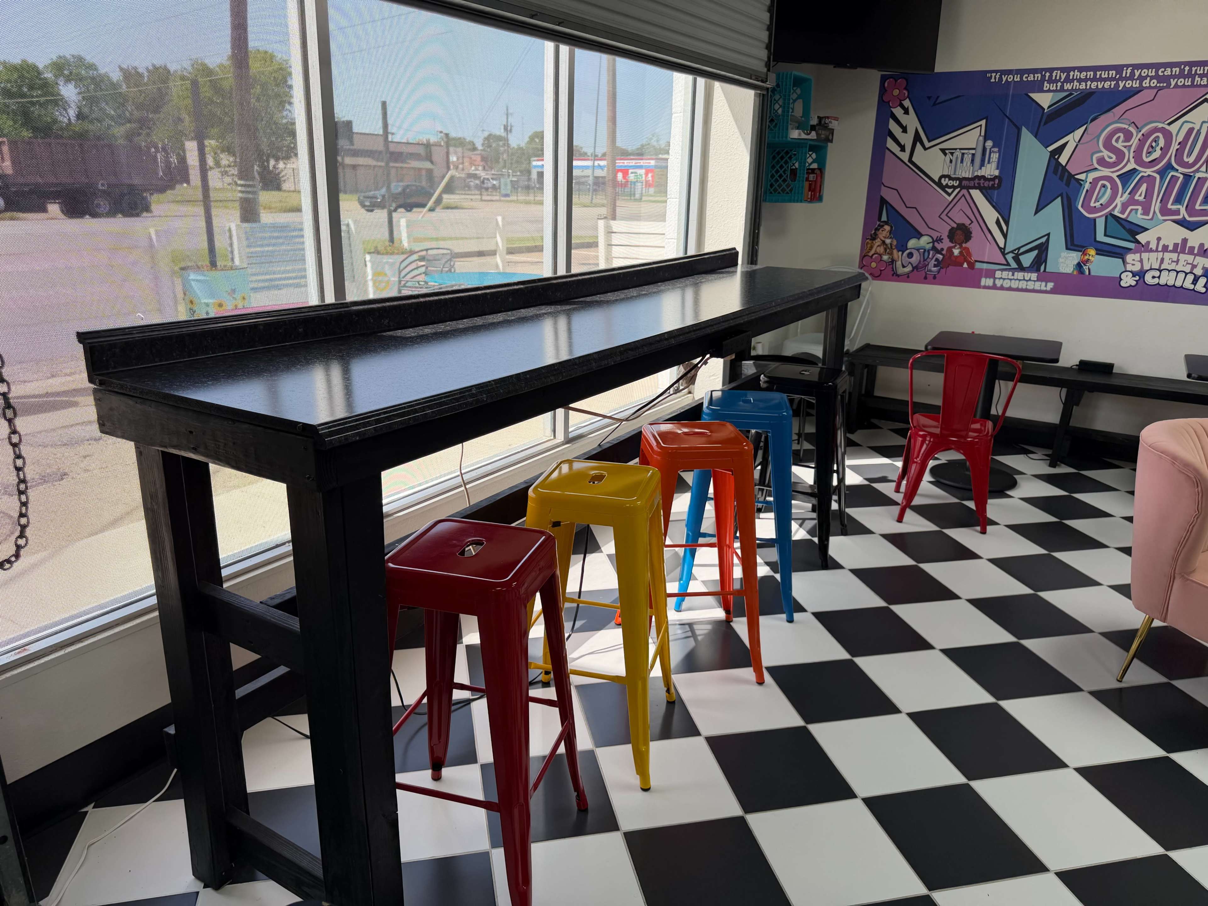 The image shows a tall black bar table with colorful metal stools in a brightly lit room featuring a checkerboard floor and large windows.
