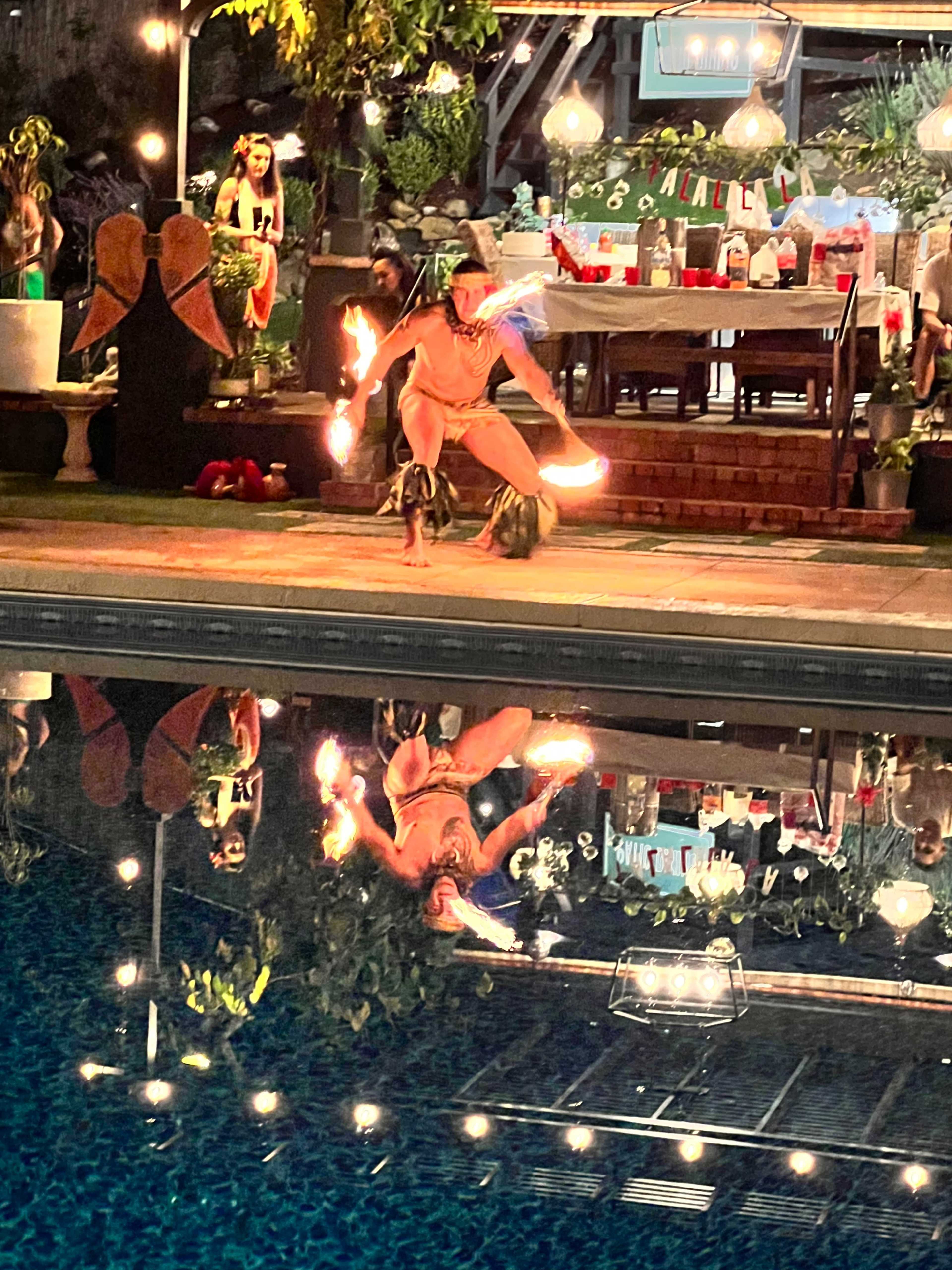 A performer is dancing with fire props by a poolside, reflecting in the water, while a festive gathering is set up in the background.