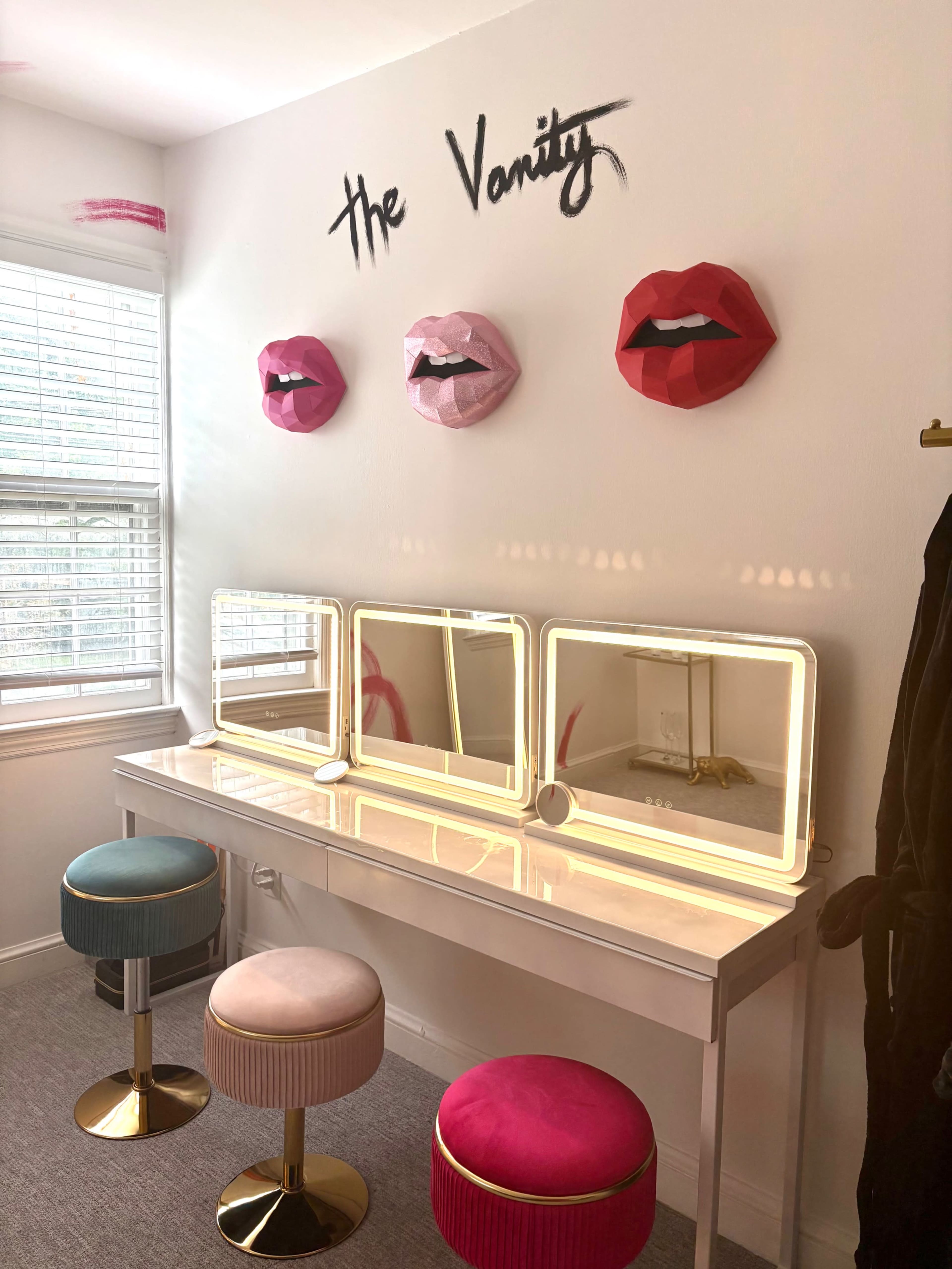 The image shows a well-lit vanity area with three decorative lips on the wall and a white table featuring three mirrors, two stools in pastel colors, and a plush pink seat.