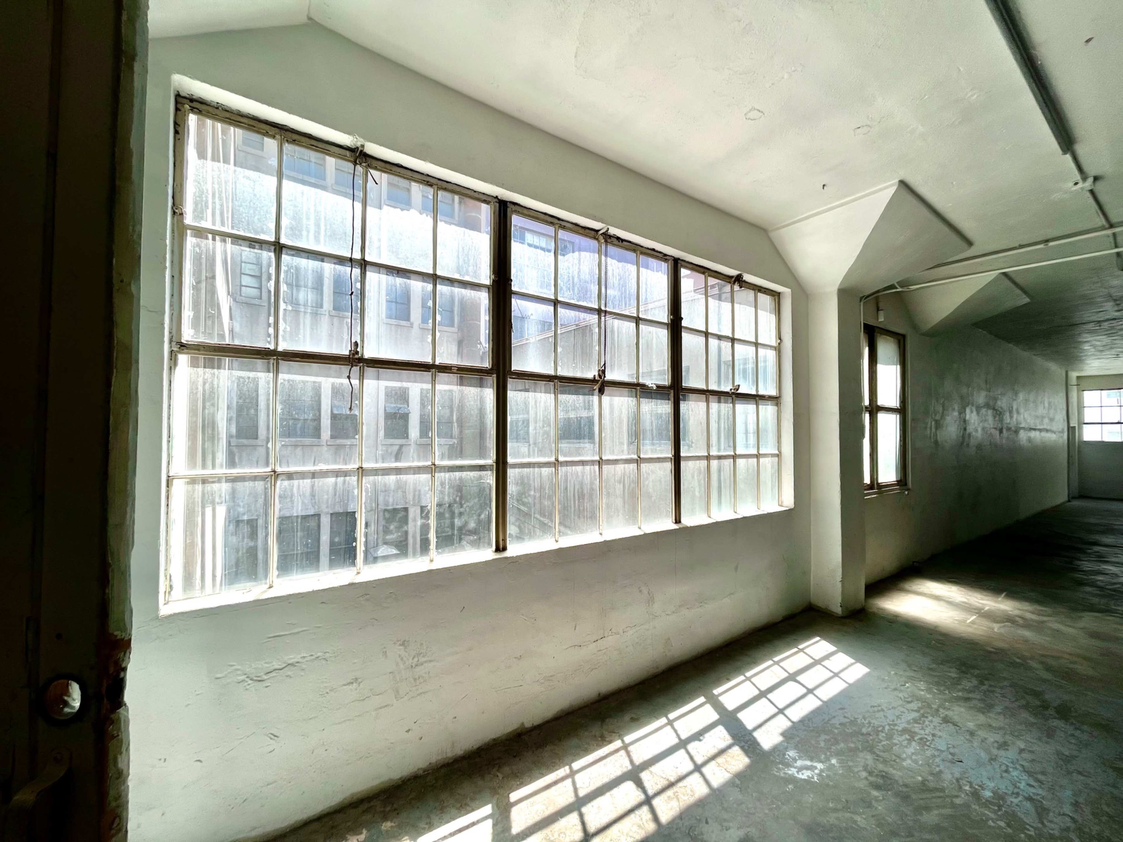 A large, multi-pane window allows sunlight to filter into a blank, spacious hallway with concrete flooring.