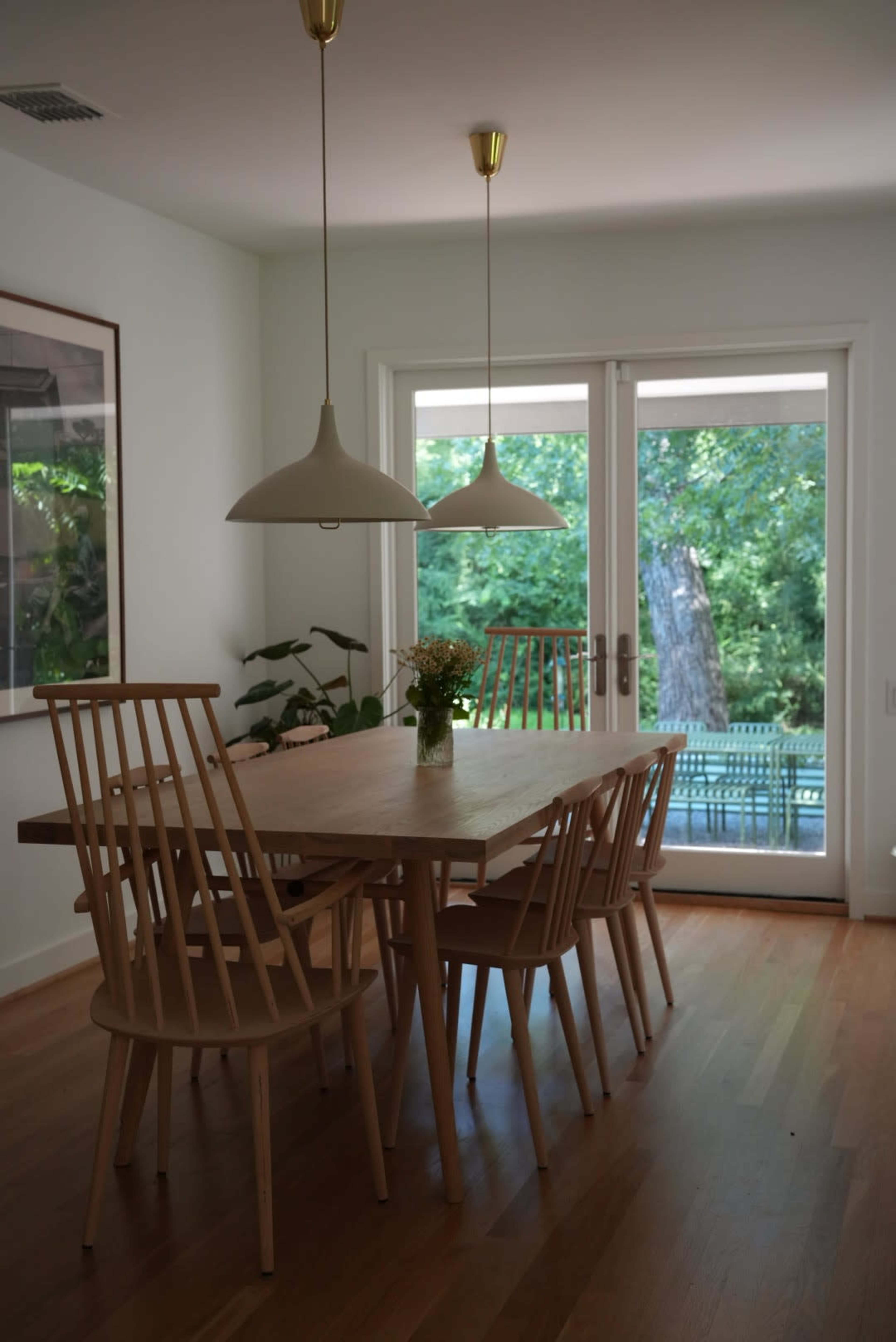 A wooden dining table surrounded by six chairs is illuminated by two pendant lights, with a view of a deck and greenery visible through the sliding glass doors.