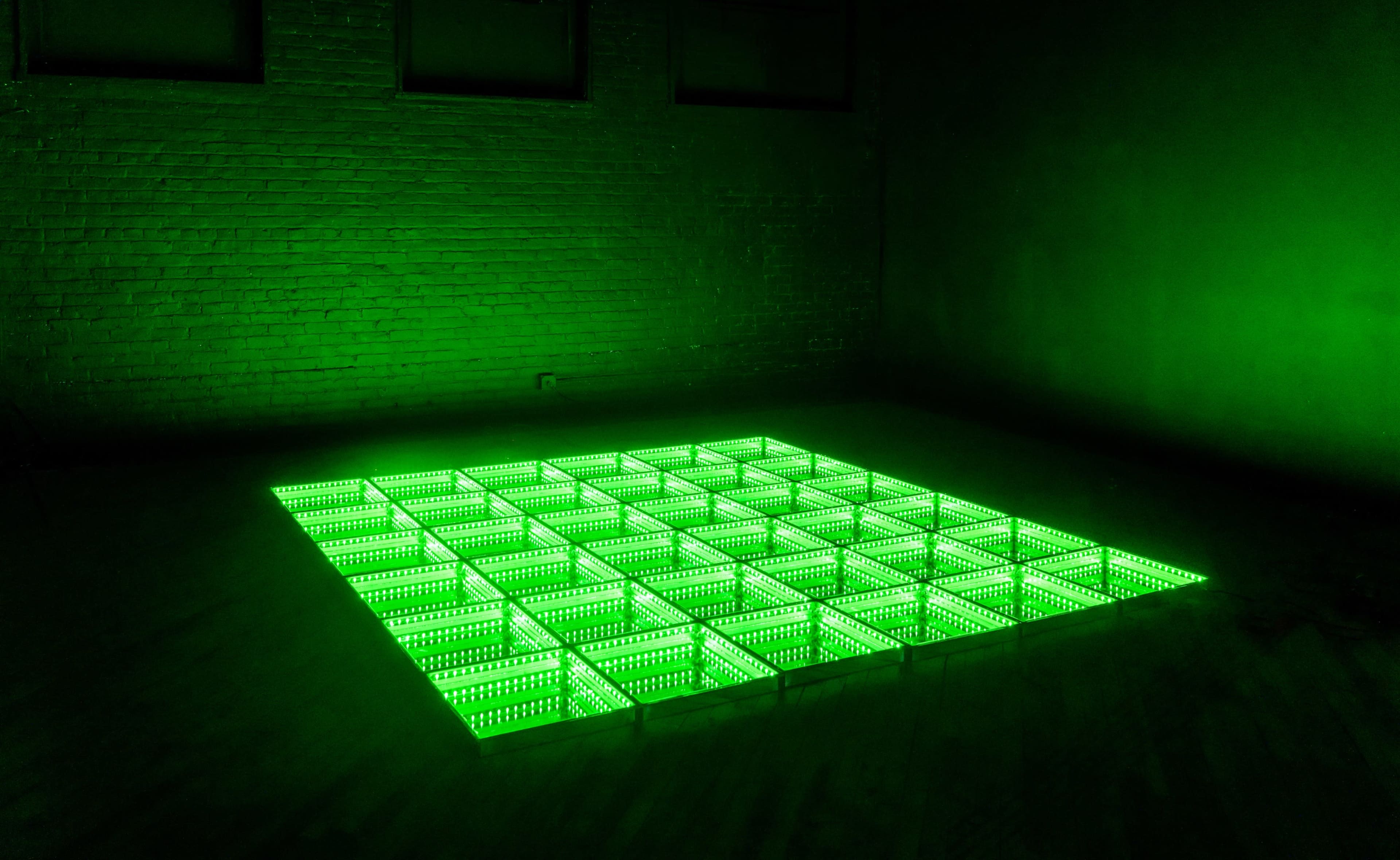 A grid of illuminated green lights arranged on the floor in a dimly lit room with brick walls.