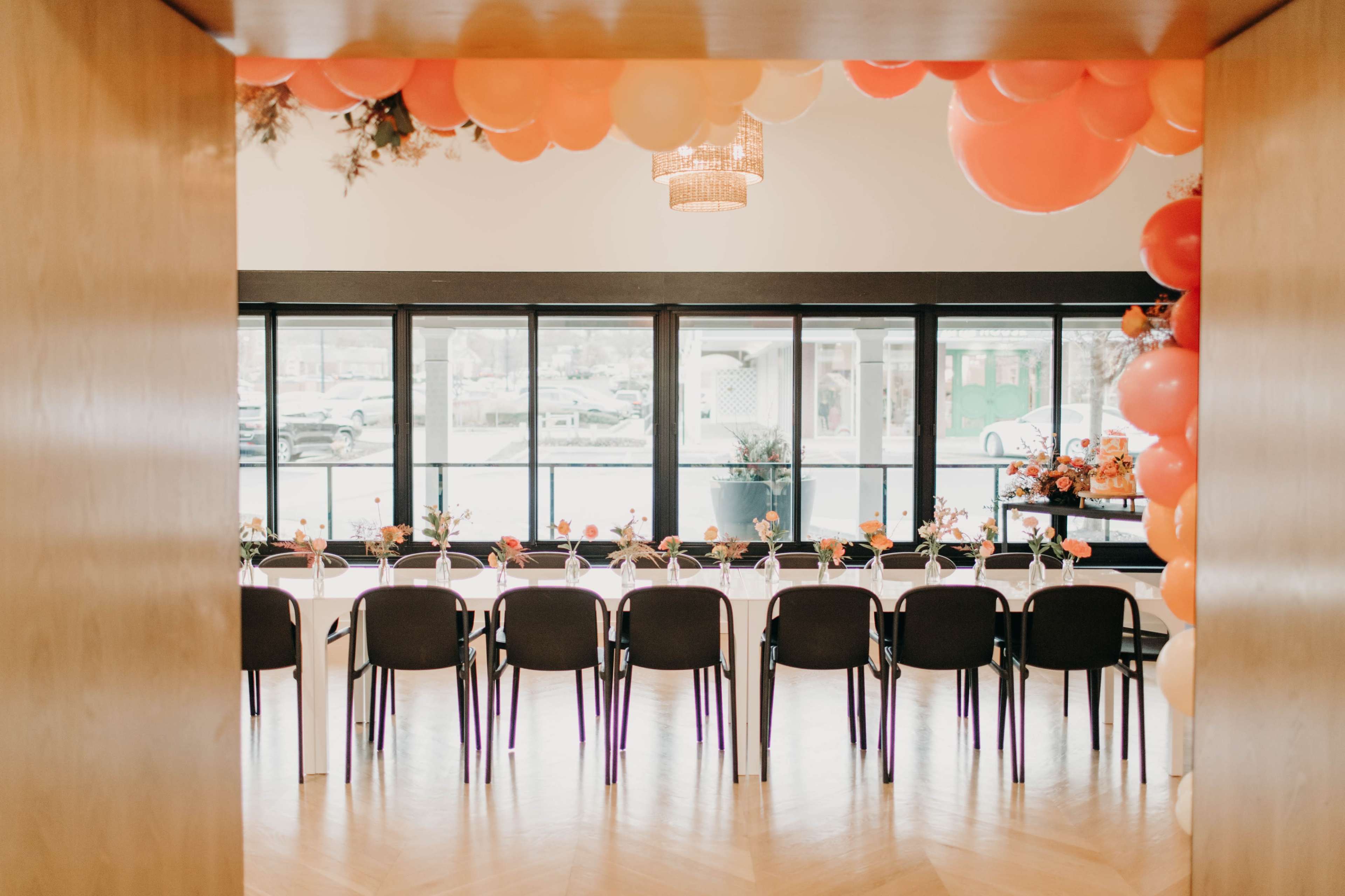 A dining room features a long table set with flowers and black chairs, surrounded by large orange and pink balloons, with large windows providing a view outside.