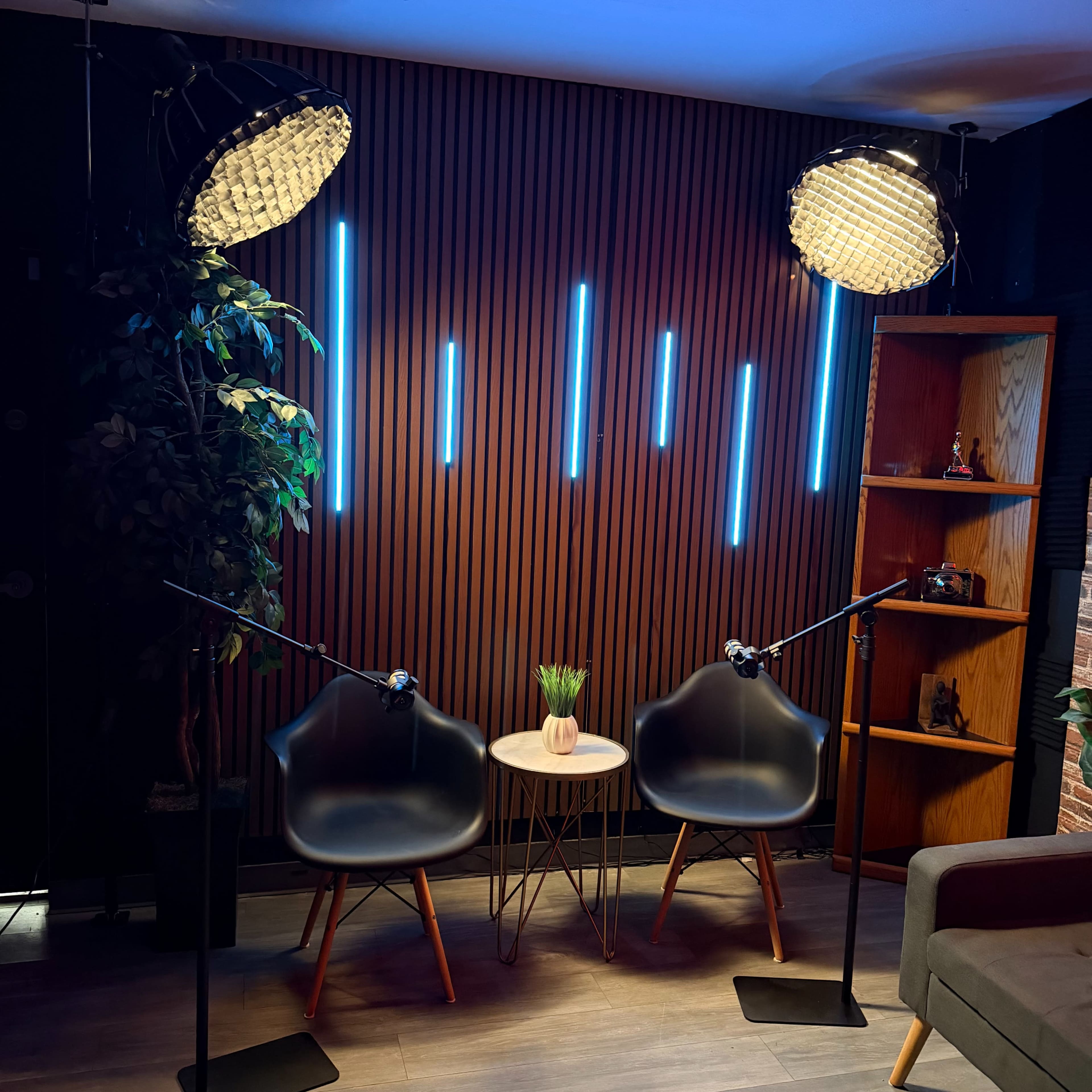 The image shows a modern interview setup with two black chairs, a small round table, and two microphones positioned in front of a wooden wall illuminated by blue LED lights.